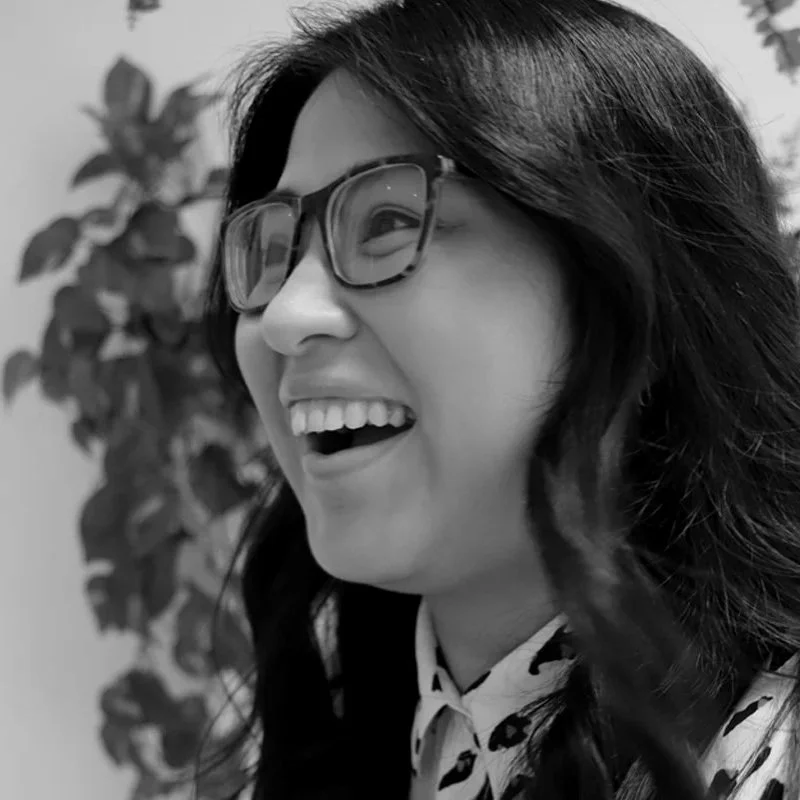 A woman with long dark hair, wearing glasses and a patterned shirt, is laughing or smiling and looking to her right, with a leafy plant in the background.