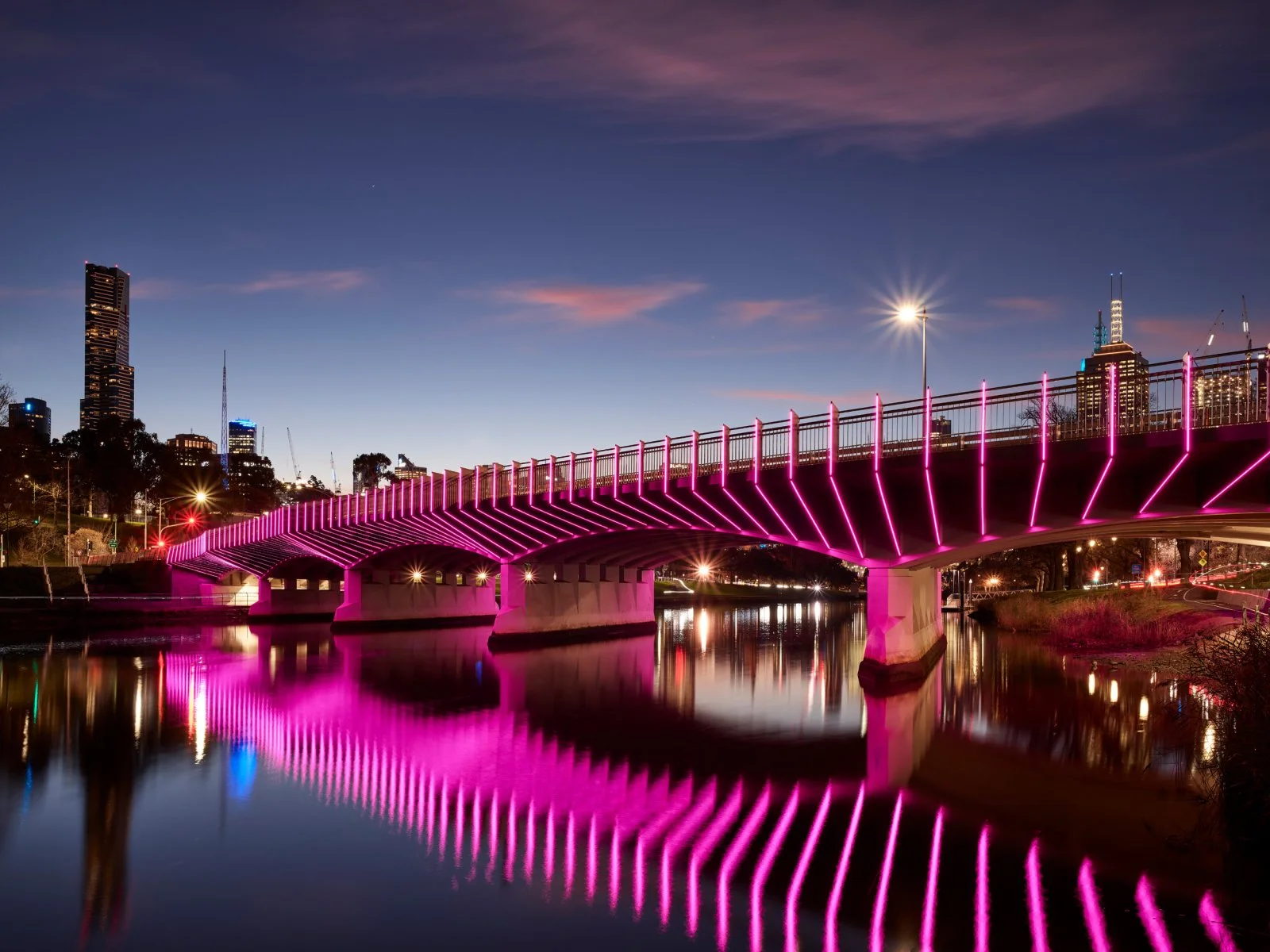A nighttime view of a city skyline with a bridge illuminated with pink neon lights spanning over a river, with reflections in the water and skyscrapers in the background.