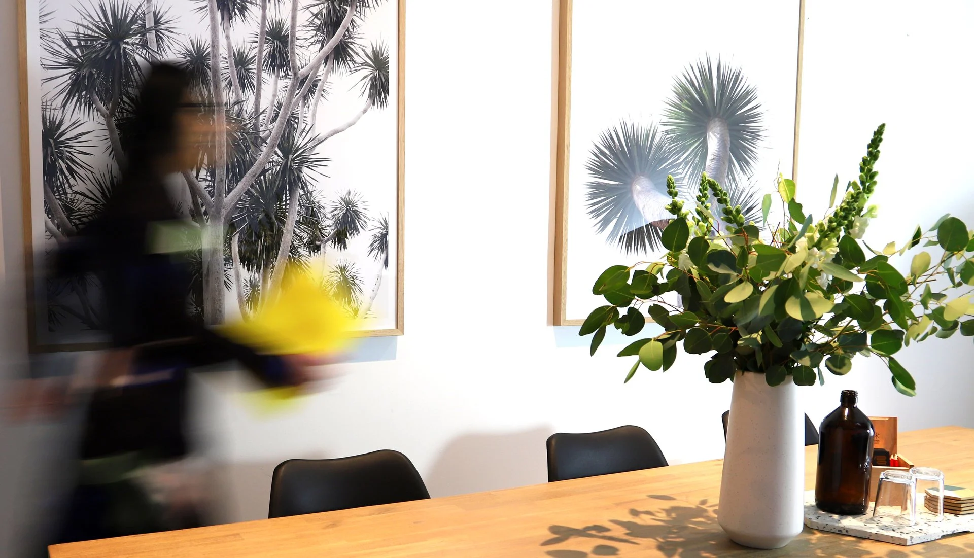 A large white vase filled with green foliage and white flowers on a wooden table, with a black bottle, glassware, and a coaster nearby. Two black and white framed botanical prints of palm trees hang on the wall in the background.