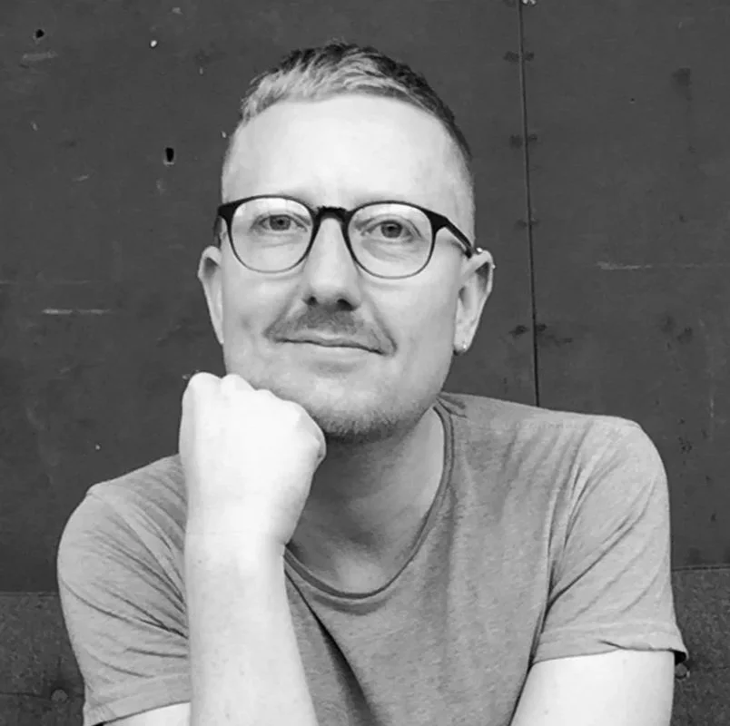 A man with glasses and a short haircut, wearing a t-shirt, sits with his hand resting against his chin, smiling slightly at the camera against a plain dark background.