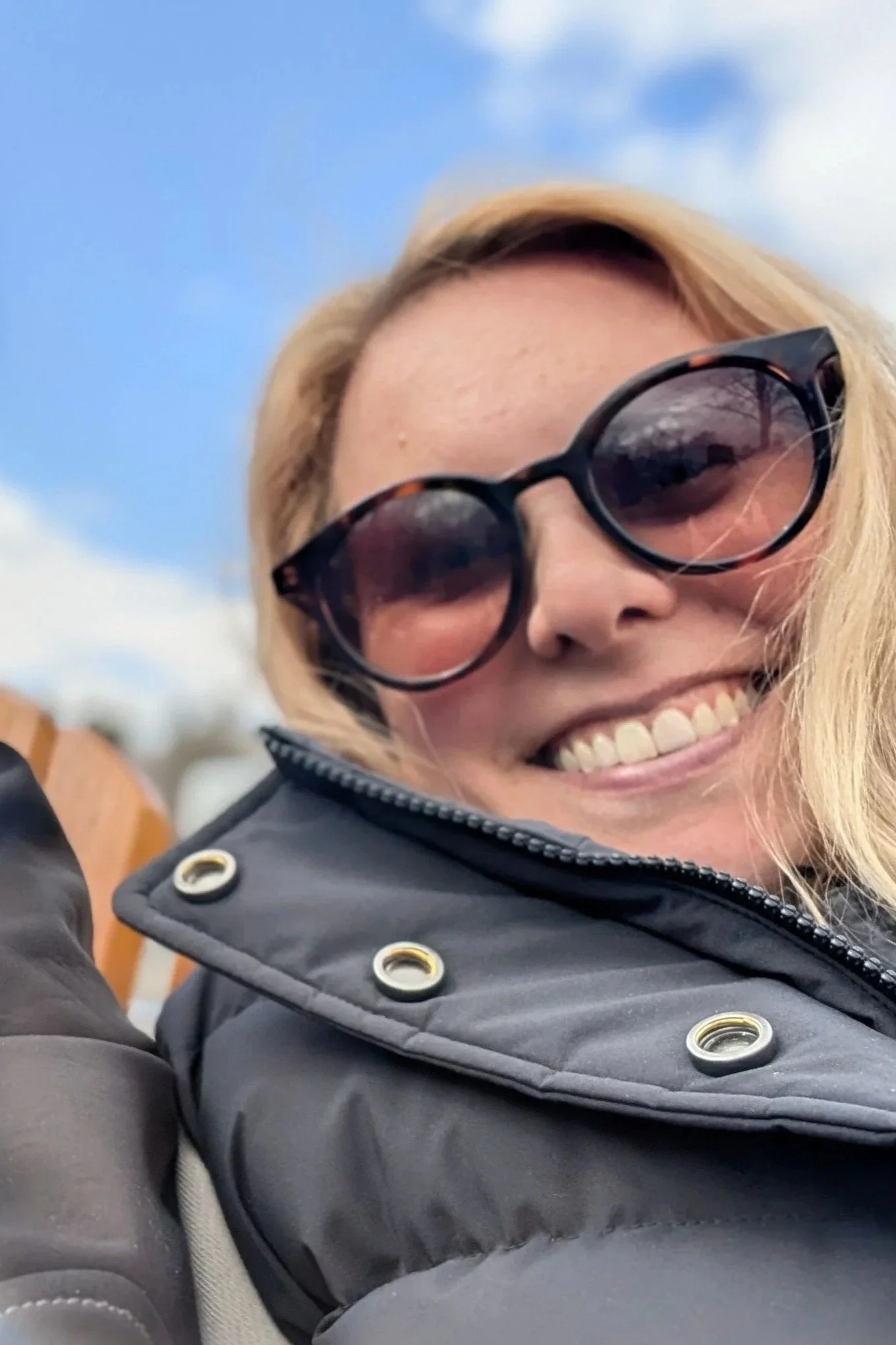 Une femme portant des lunettes de soleil sourit, avec un ciel bleu en arrière-plan.