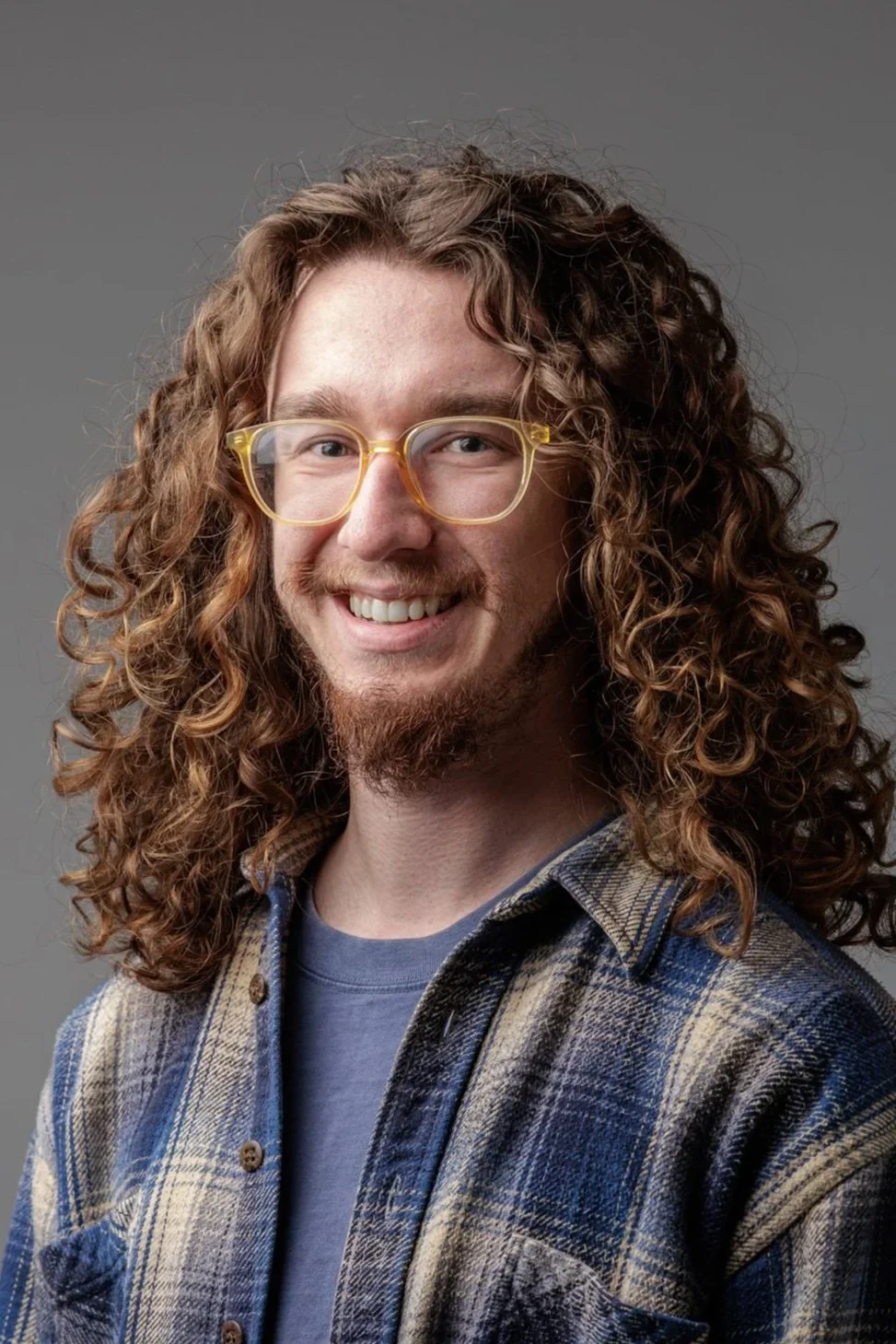 Portrait of a man with long curly hair, glasses, and a beard, smiling, wearing a plaid shirt and a blue T-shirt, against a gray background.