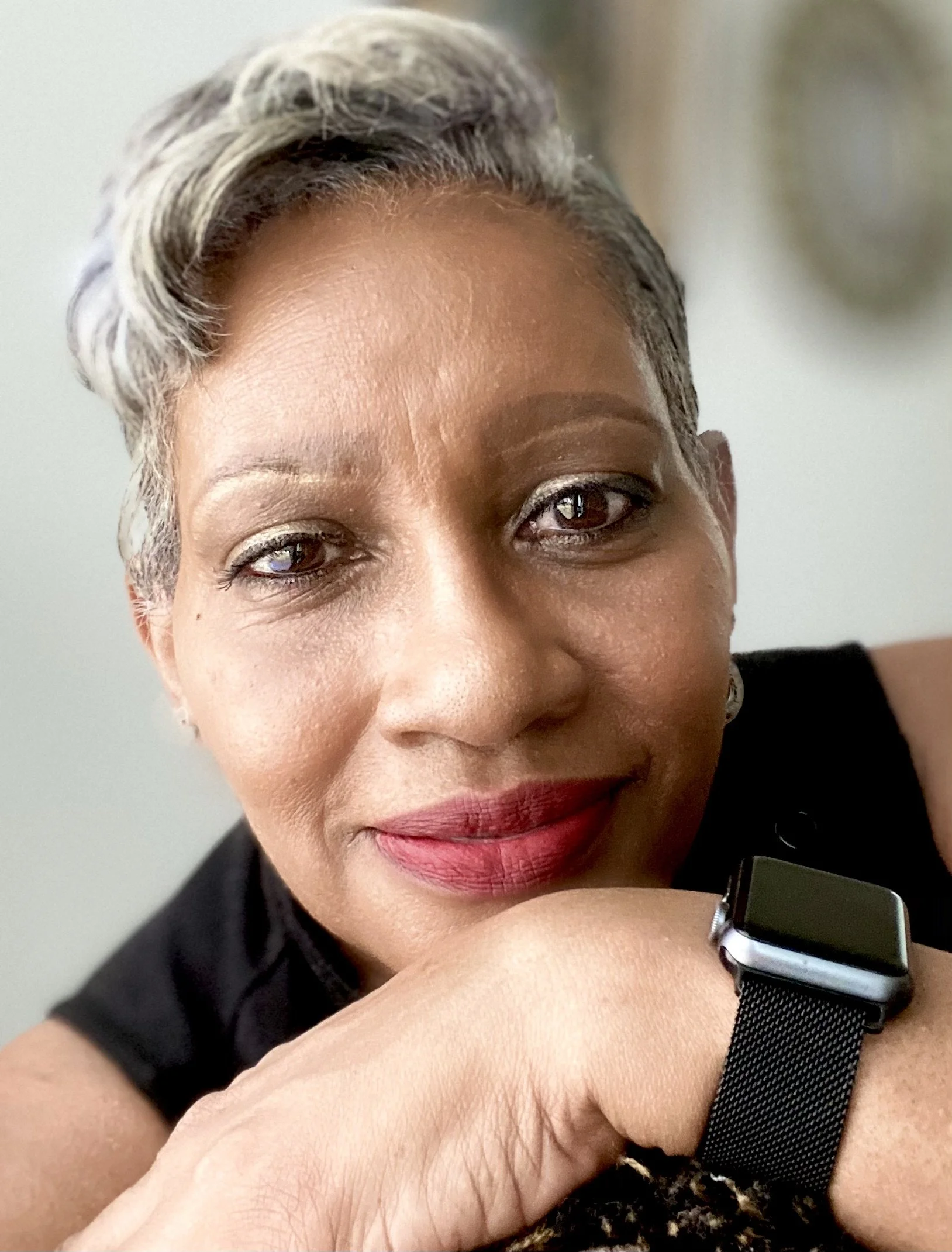 Close-up of a smiling woman with short, gray and black hair, wearing a black sleeveless top, red lipstick, and an Apple Watch on her wrist.