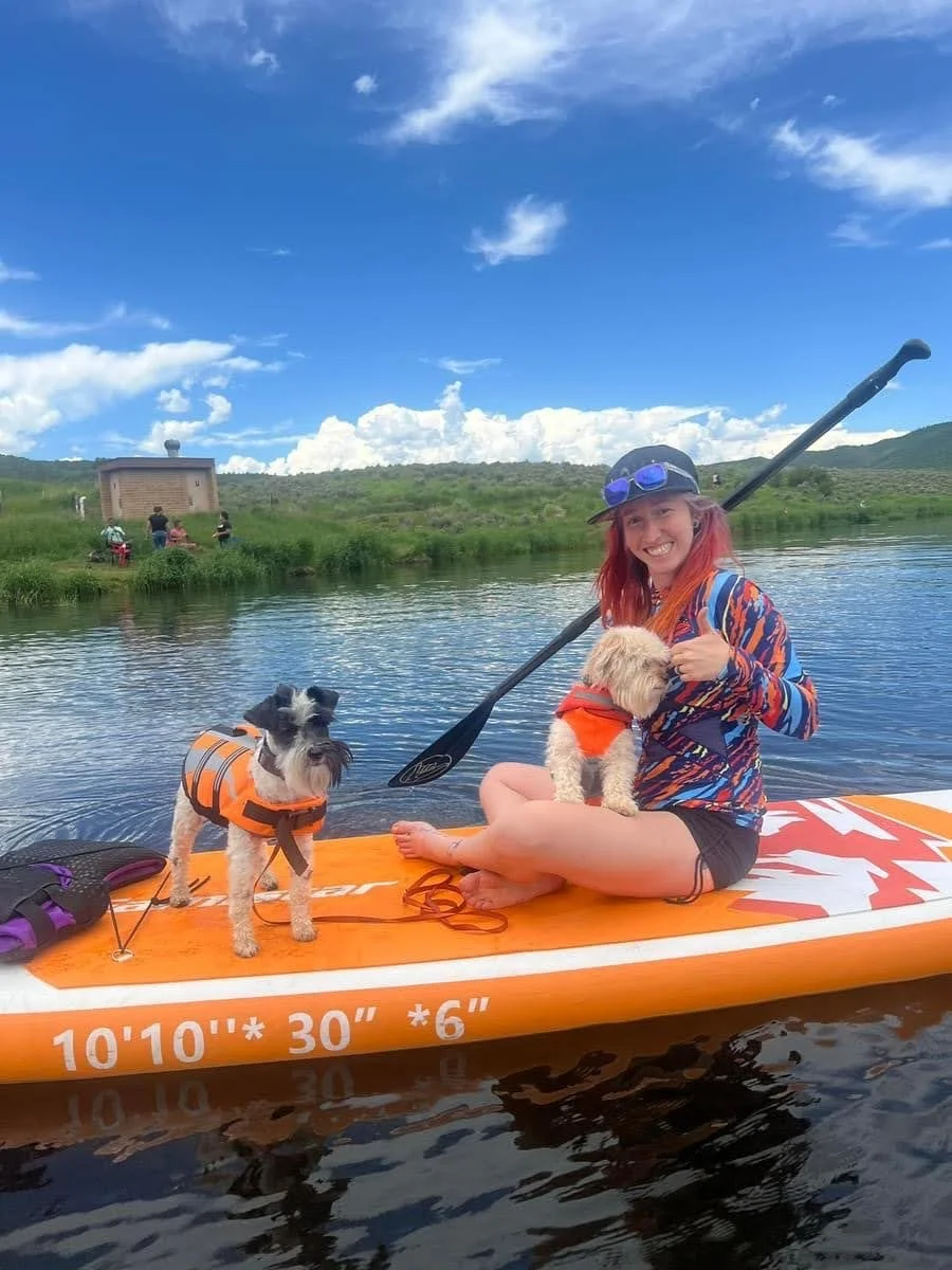 A woman with red hair, wearing a cap and sunglasses, sitting on an orange paddleboard with two small dogs, one sitting on her lap and the other standing next to her, on a calm river with a grassy shore and a sky with scattered clouds in the background.