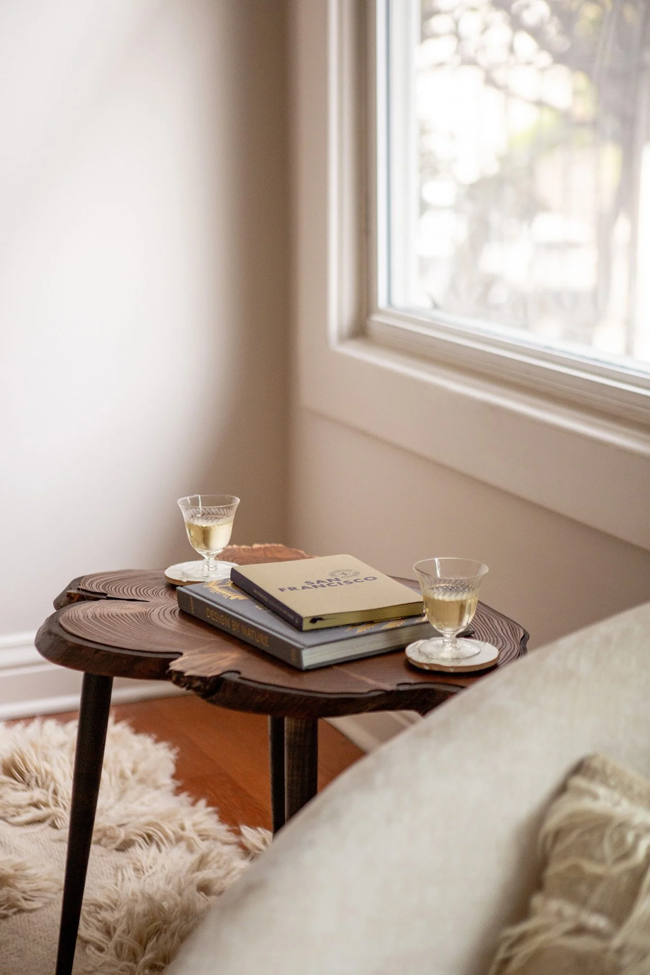 Light-filled living area detail at Pine Street Retreat showing a wood side table with wine glasses and San Francisco books beside softly upholstered hair with textural vintage shag pillow in a tonal neutral hue.