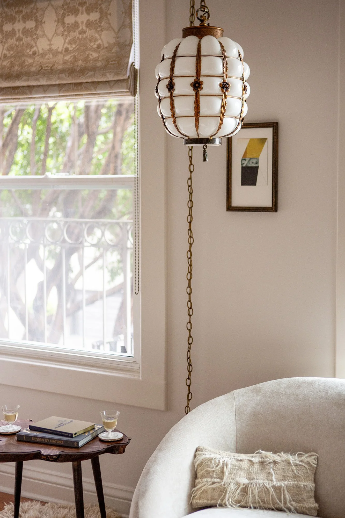 Vintage milk glass pendant light with brass chain hanging beside a curved white armchair, wood side table with books and wine glasses, and tree views through an iron-railed window at Pine Street Retreat in Nob Hill San Francisco