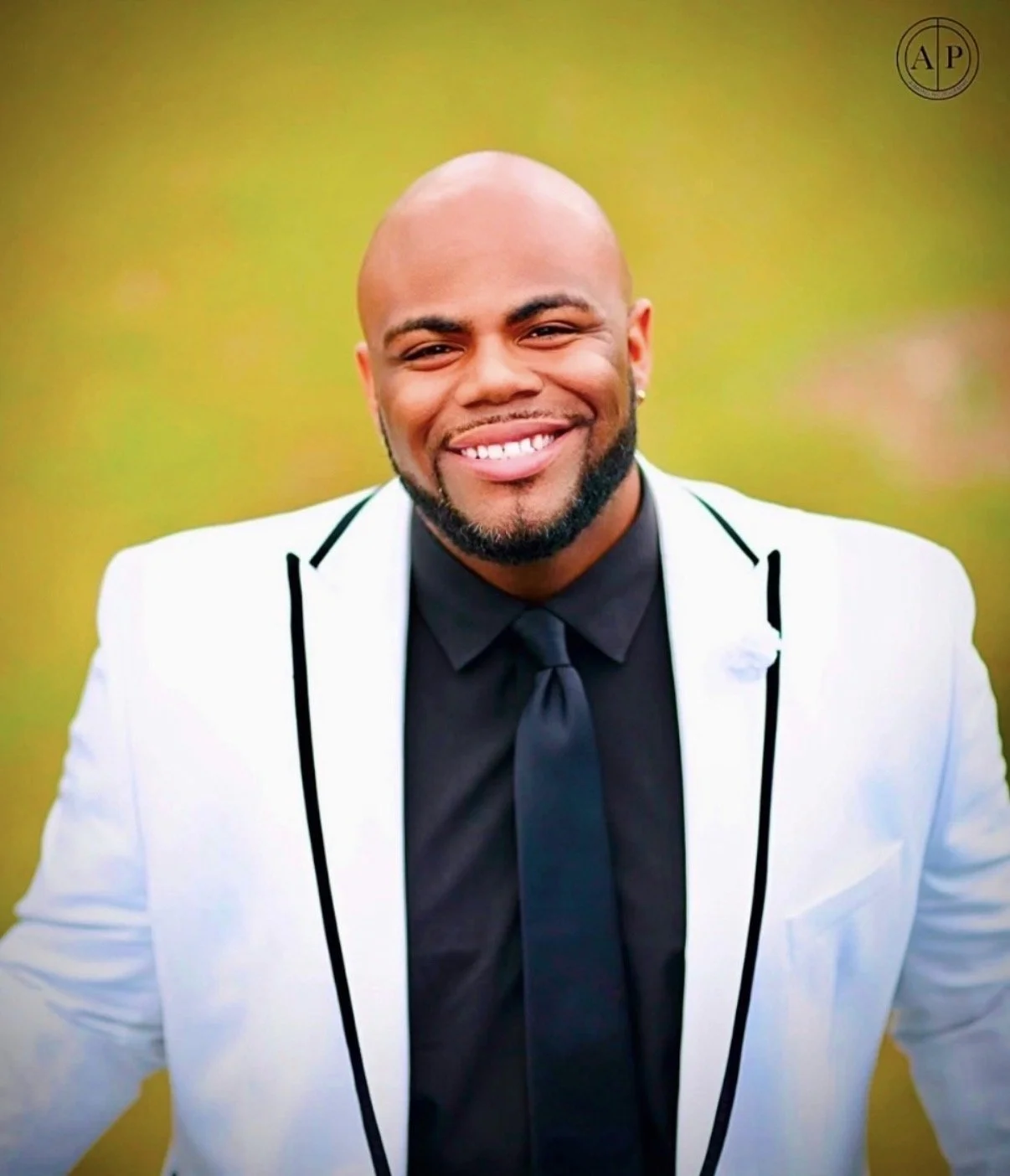 A smiling man with a bald head and beard, wearing a white blazer with black trim, a black shirt, and a black tie, standing outdoors with a blurred green background.