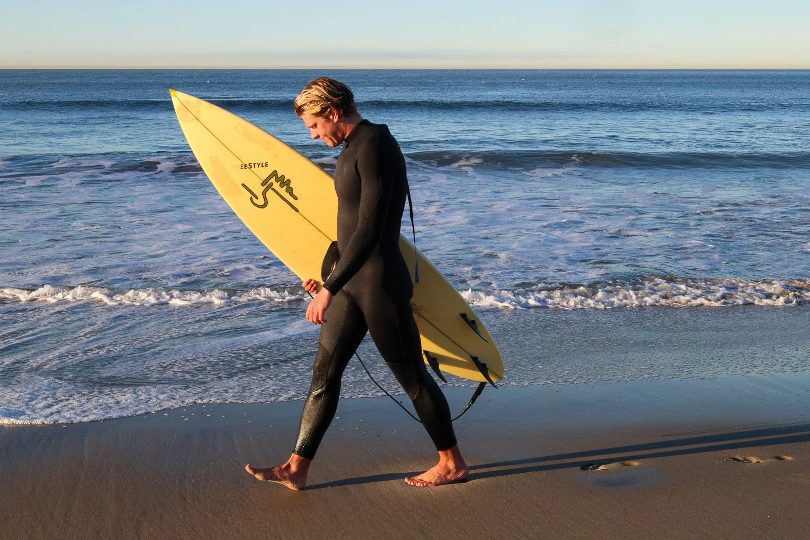 A person in a black wetsuit holding a yellow surfboard walking on the beach near the ocean at sunset.