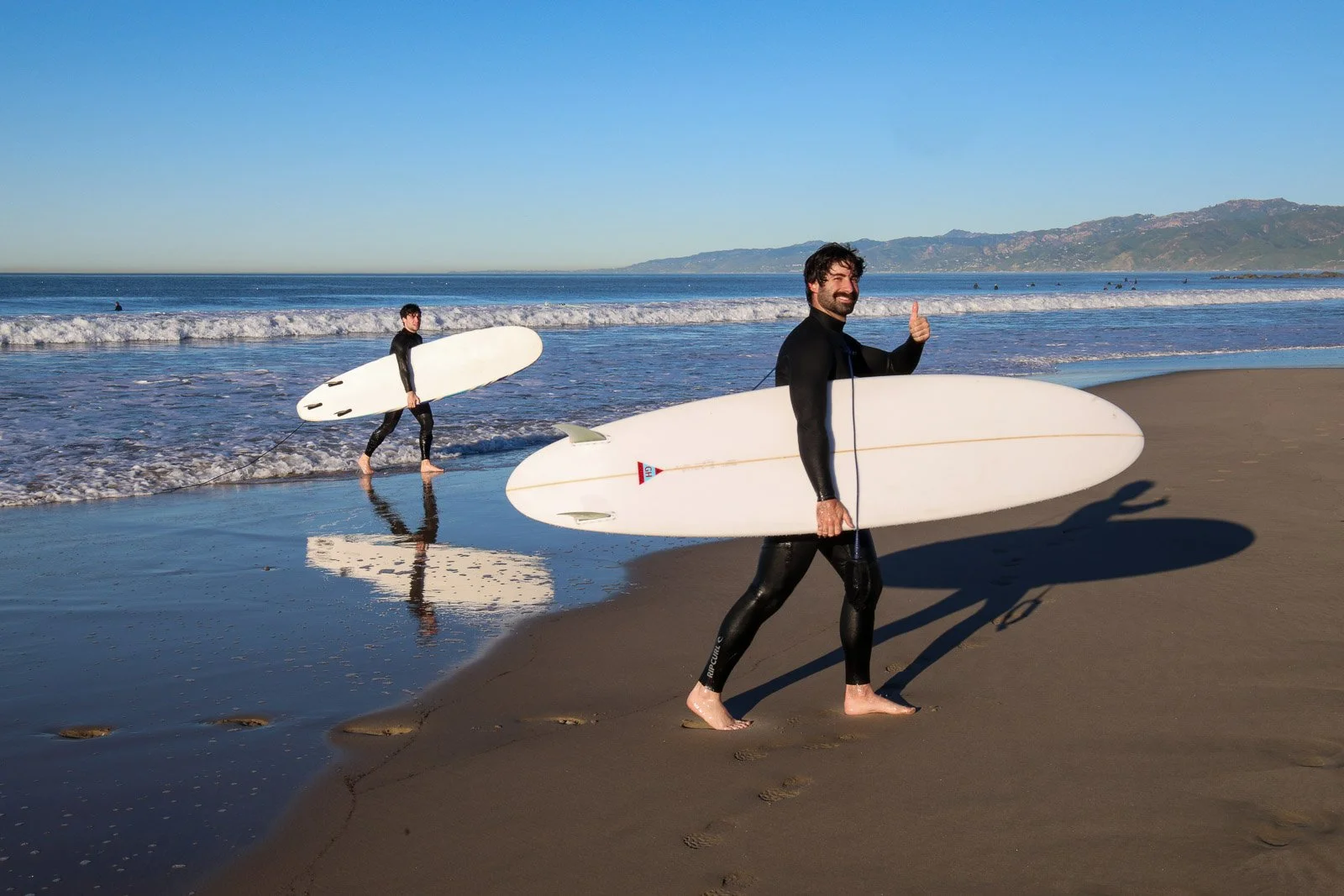 Two surfers in wetsuits walking on the beach with surfboards, one giving a thumbs-up gesture.