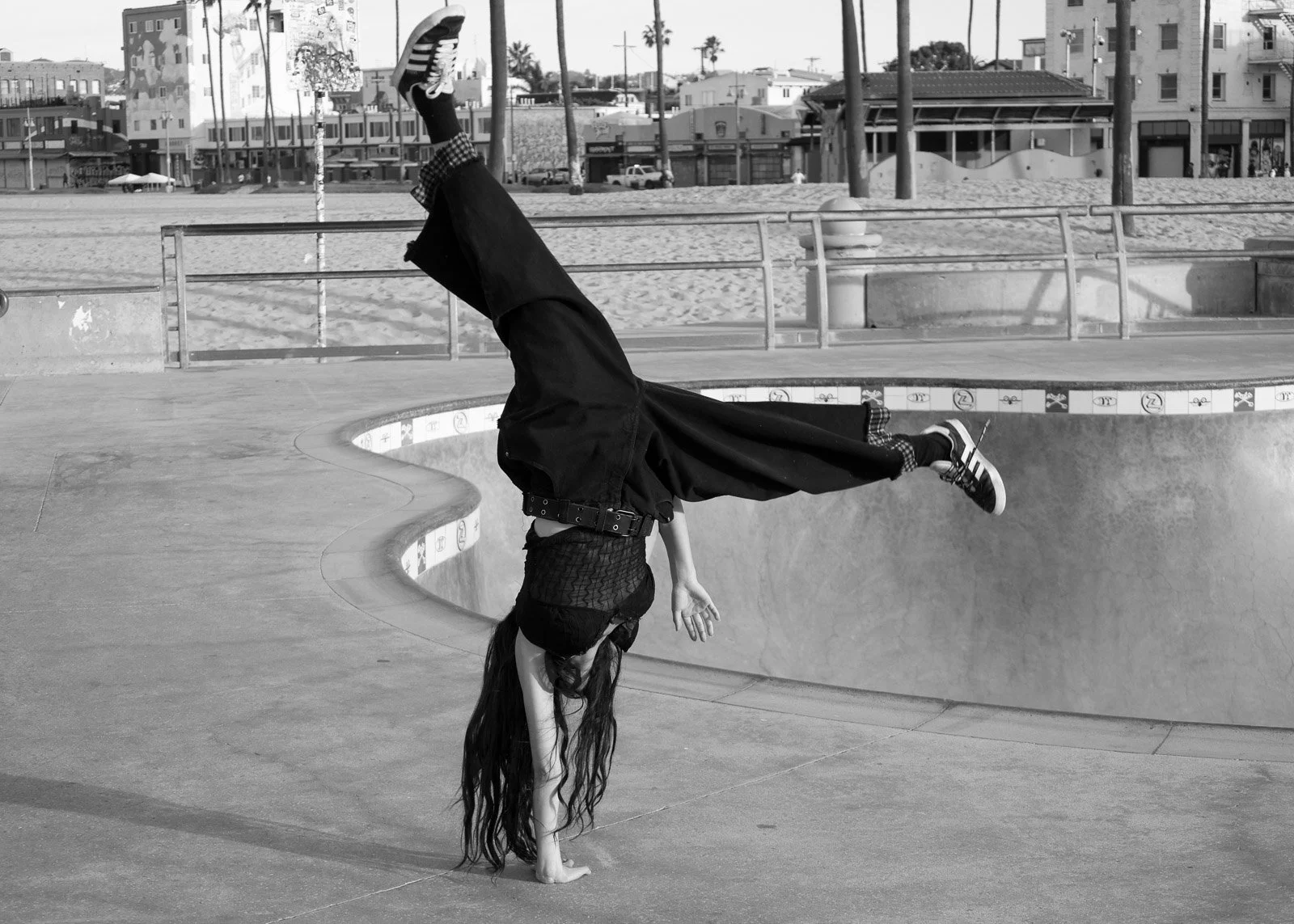 A person with long hair performing a handstand on the edge of an empty skate park bowl at the beach, with buildings, palm trees, and a clear sky in the background.