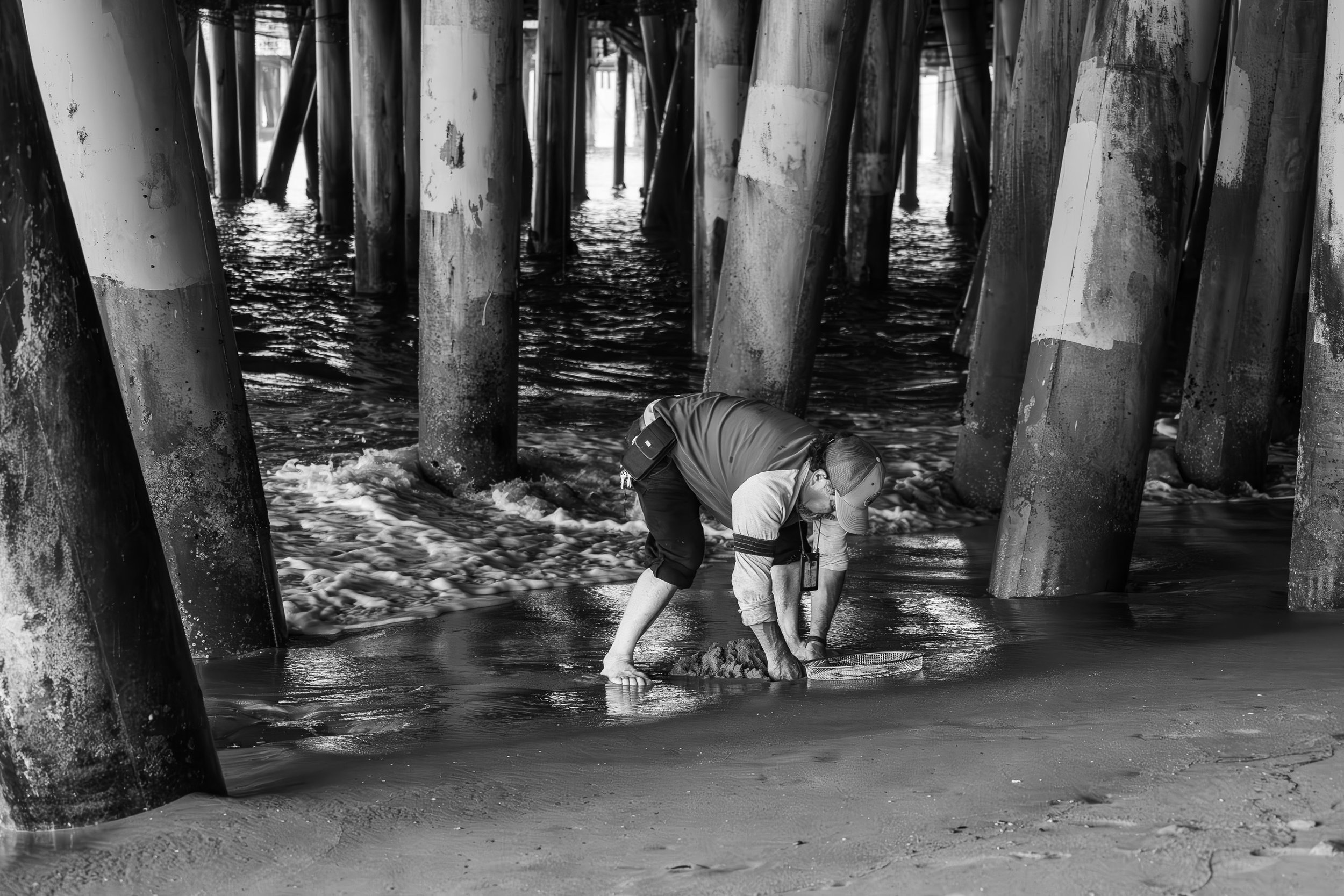 A person digging in the sand near the water under a pier with large support beams.