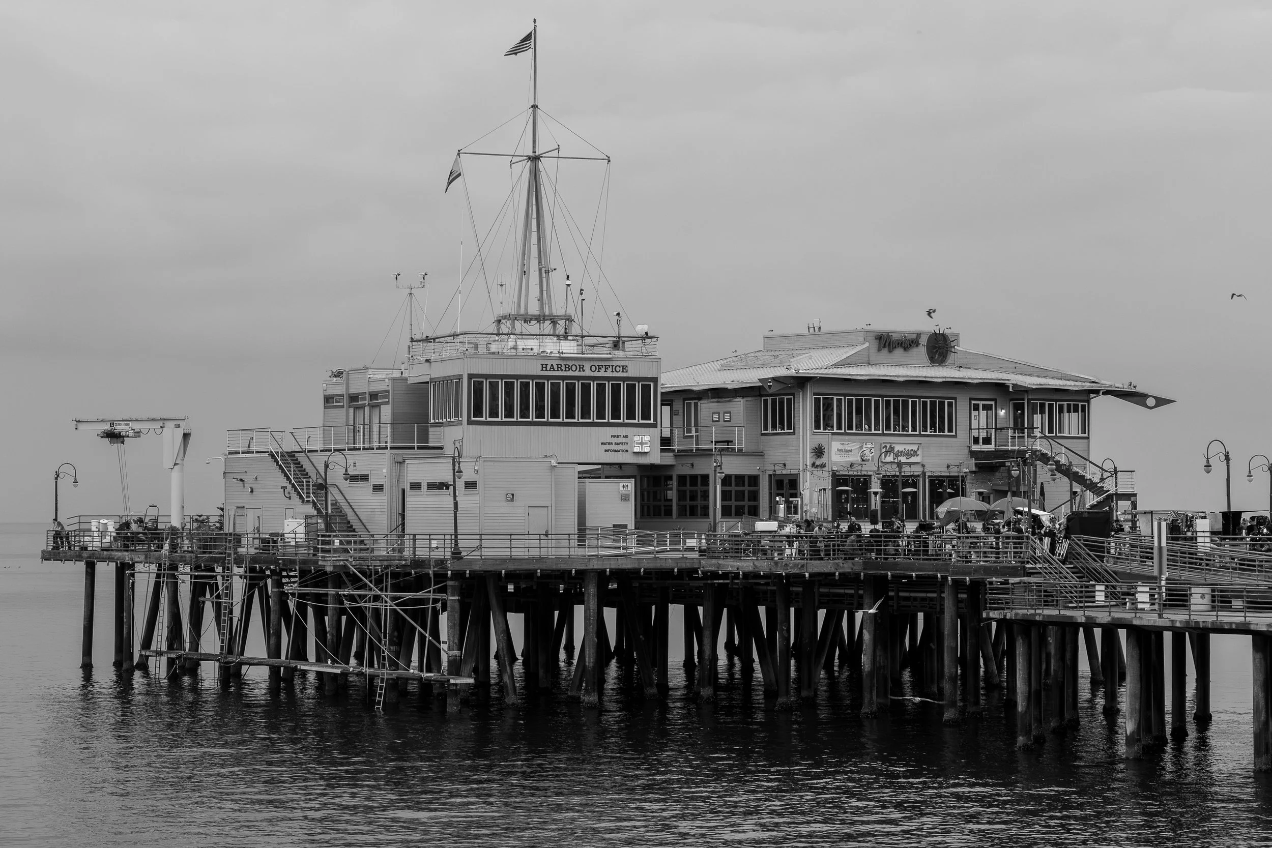 A black and white photo of a pier extending into the water with a building on it. The building has signs reading 'Harbor Office' and 'Margaritaville,' and features a ship's mast, windows, balconies, and outdoor seating, with several people and umbrel