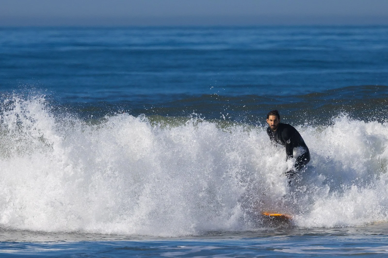 A man surfing on a wave in the ocean, wearing a black wetsuit, with blue water and sky in the background.