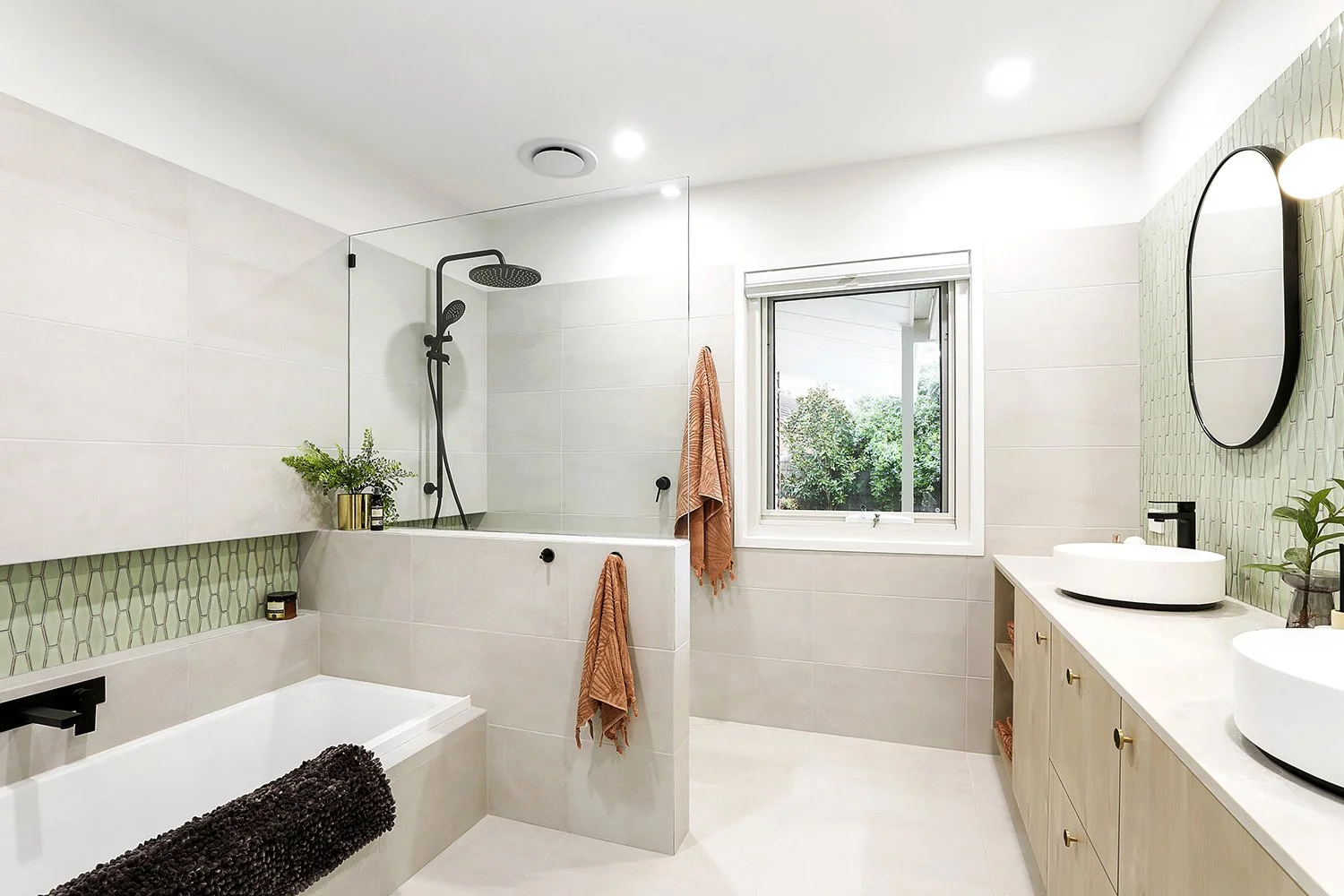 Modern bathroom with white and beige tiles, double sinks, a window, and a walk-in shower with a glass partition.