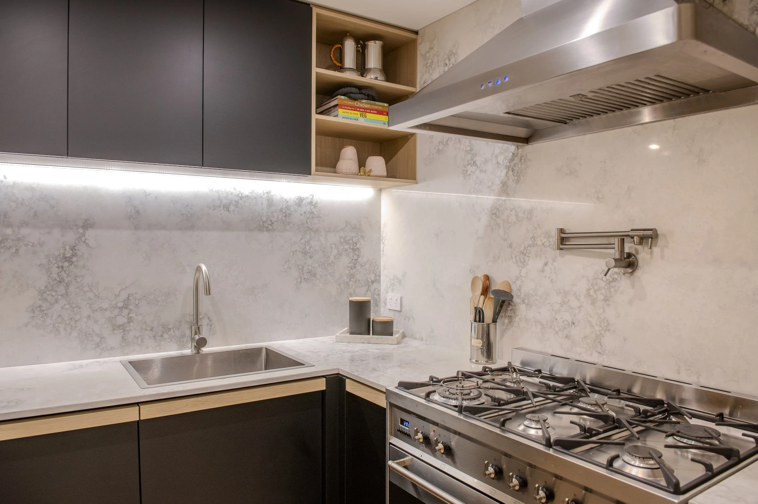Modern kitchen with black cabinets, wooden open shelves with cookbooks and decor, stainless steel range hood, marble backsplash, sink, and gas stove.