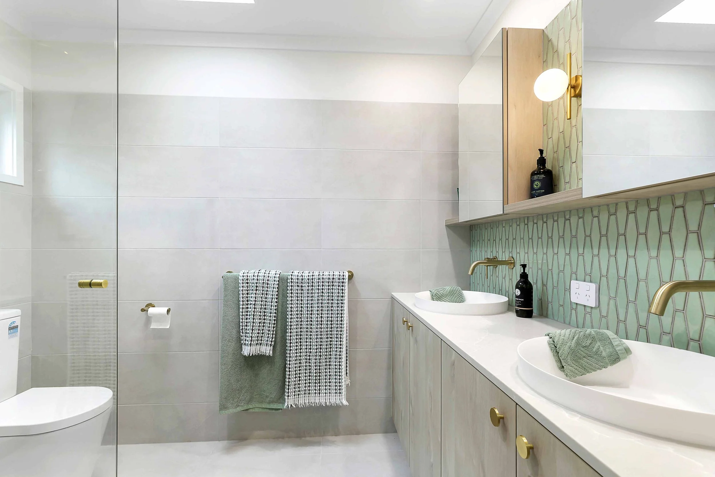 Modern bathroom with double vanity, round sinks, gold faucets, and large mirror. Towel rack with three towels, toilet in the background, and decorative tile backsplash behind the sinks.