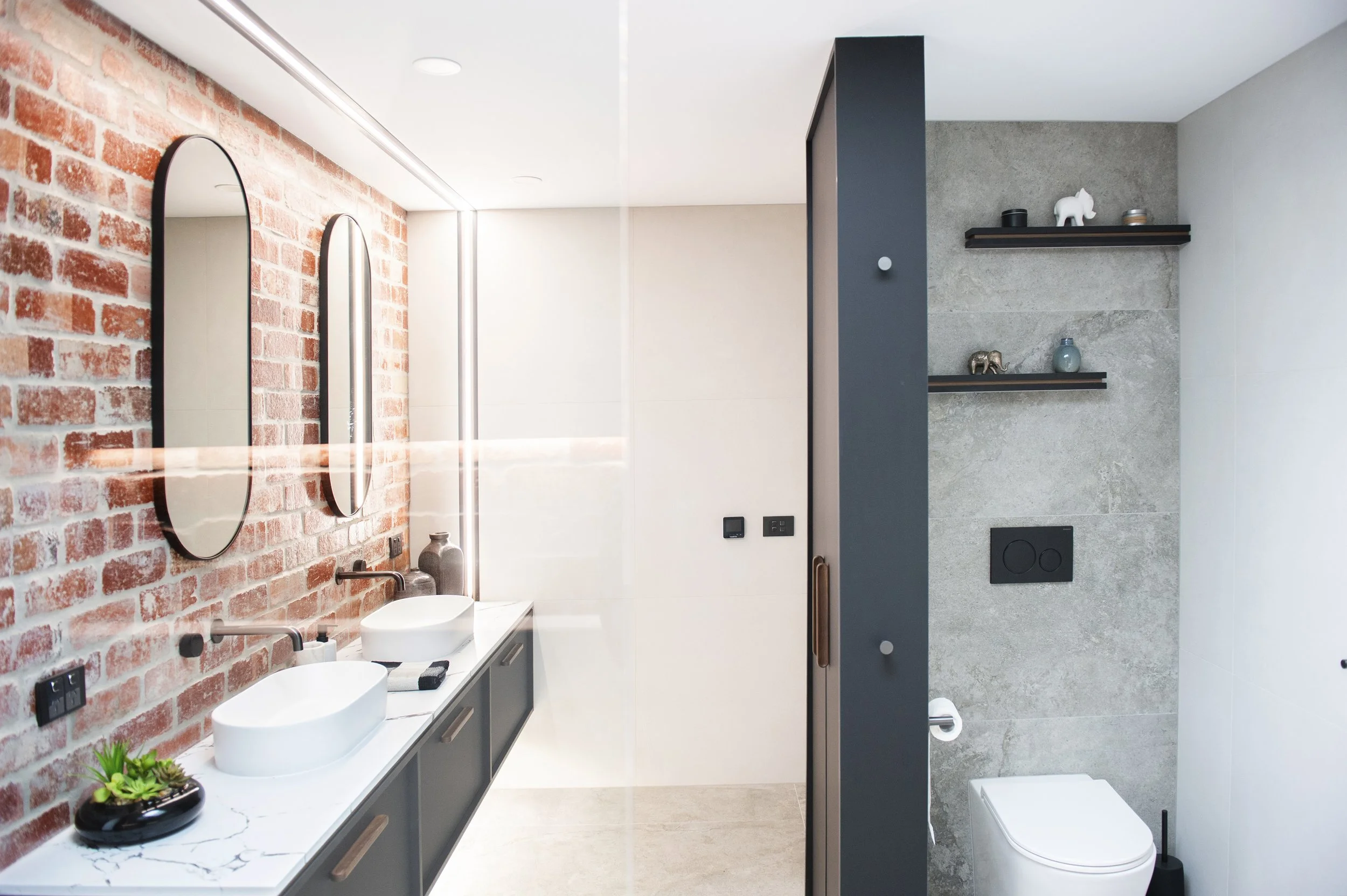 Modern bathroom with exposed brick wall, two oval mirrors, double sinks, and a planter with succulent on marble countertop. Toilet and gray shelving with decorative items on a textured wall on the right.