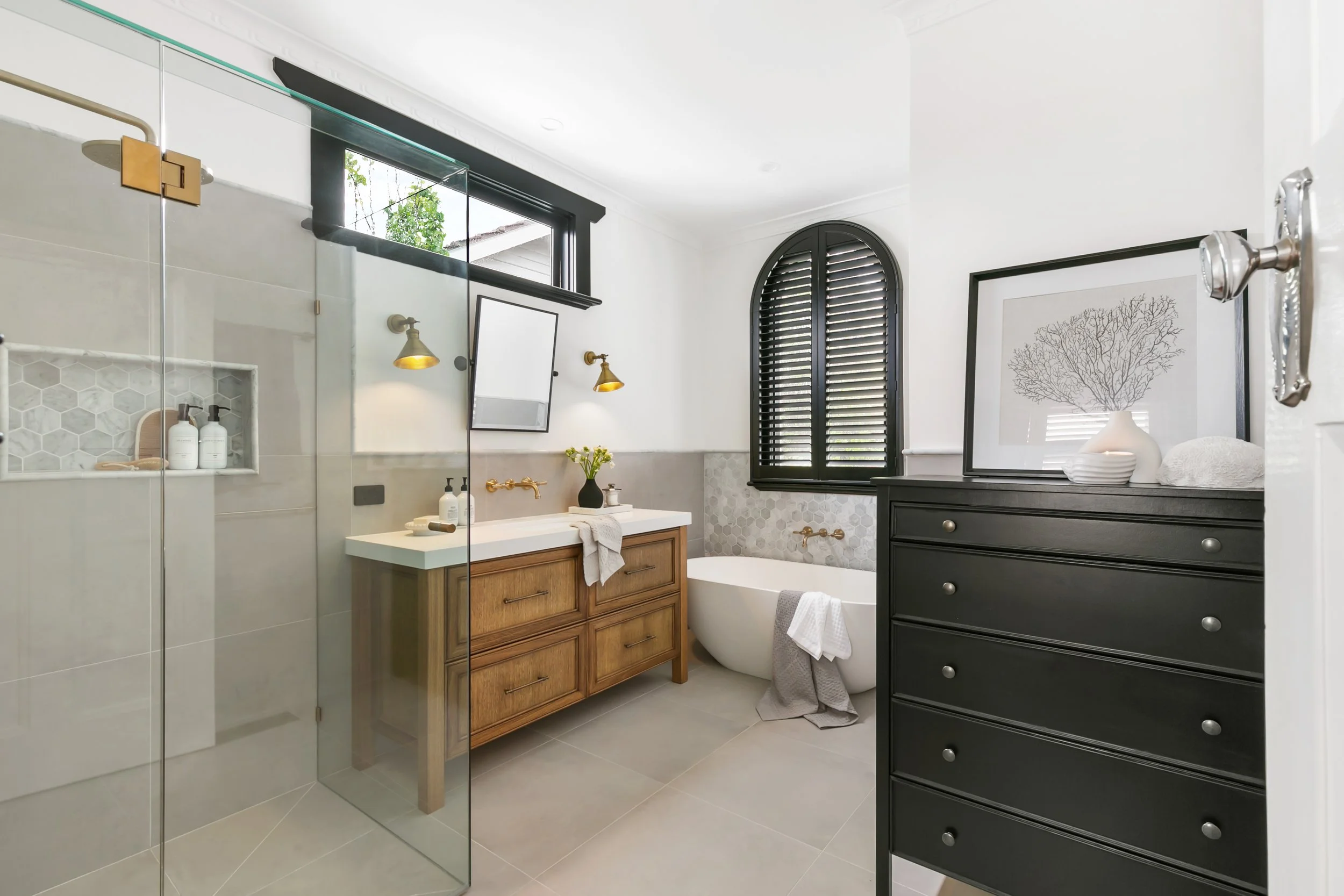 Modern bathroom with a walk-in shower, wooden vanity, bathtub, black window shutters, and black dresser topped with a framed picture and decorative vase.
