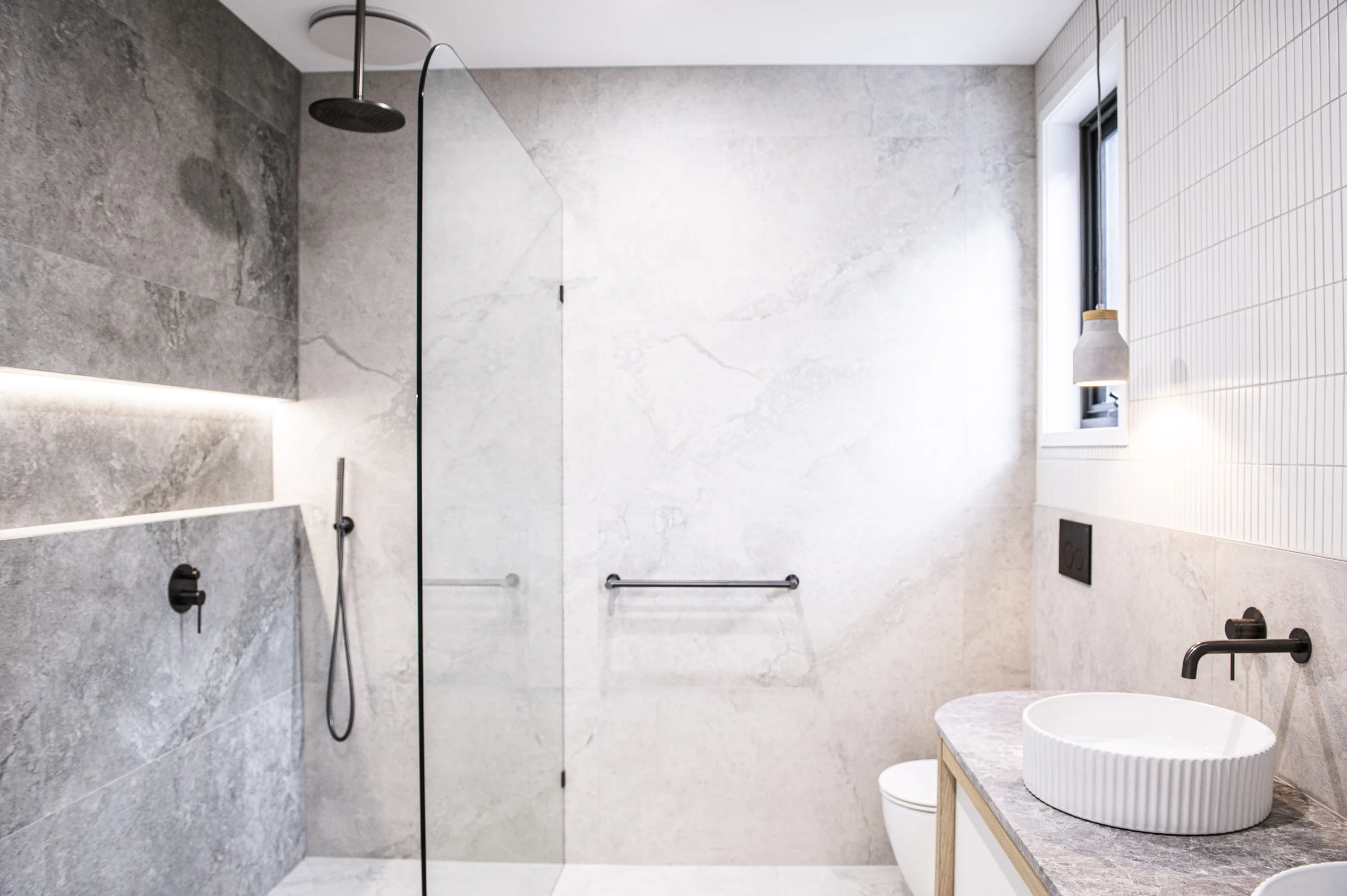 Modern bathroom with gray and white marble walls, a black rain shower, a glass shower partition, a white toilet, a circular vessel sink on a gray countertop, a small window, and minimalist black fixtures.