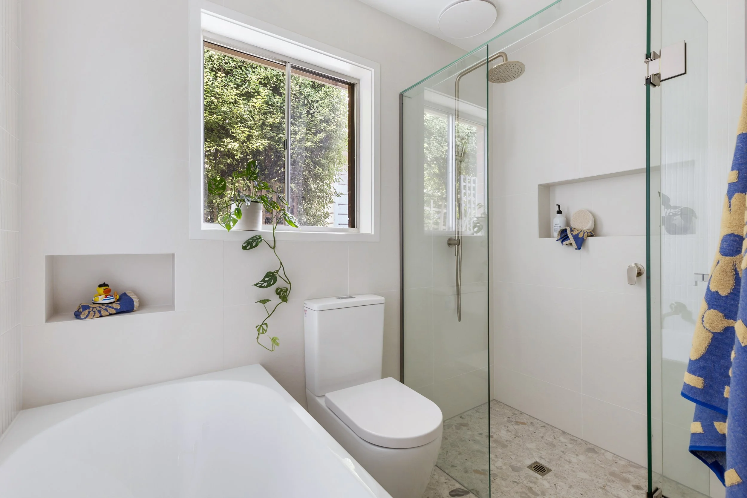 Modern white bathroom with a large window, a plant on the windowsill, a white toilet, and a walk-in glass shower with a built-in niche, soap dispenser, and a showerhead, featuring towels and a textured floor.