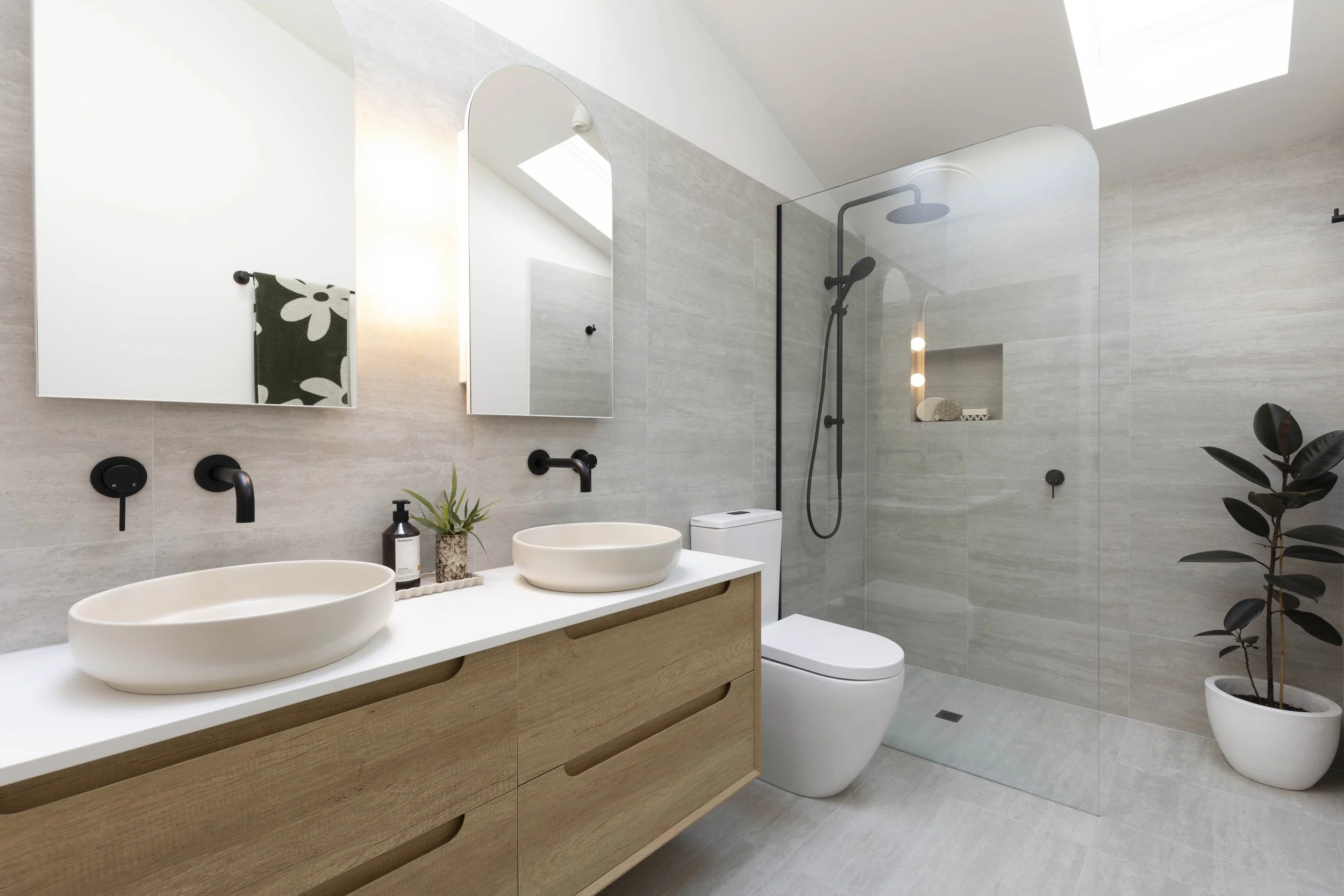 Modern bathroom with dual white vessel sinks on a wooden vanity, black fixtures, a glass shower partition, a walk-in shower with gray tiles, a skylight, and a potted plant.