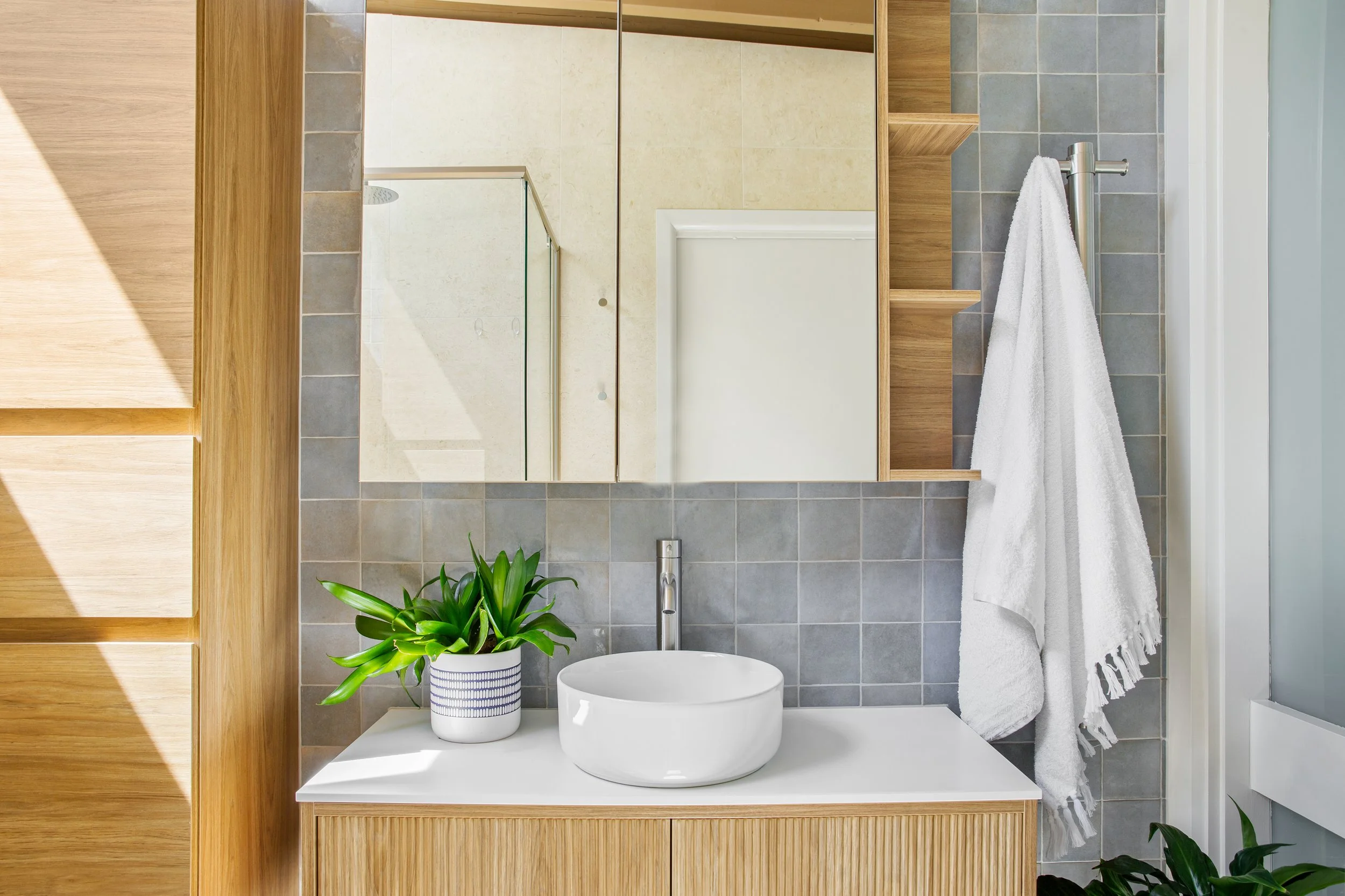 A modern bathroom vanity with a white countertop, a round vessel sink, a chrome faucet, a potted green plant, a large mirror, wooden cabinets, a white towel hanging, and a shelf with small decor items.