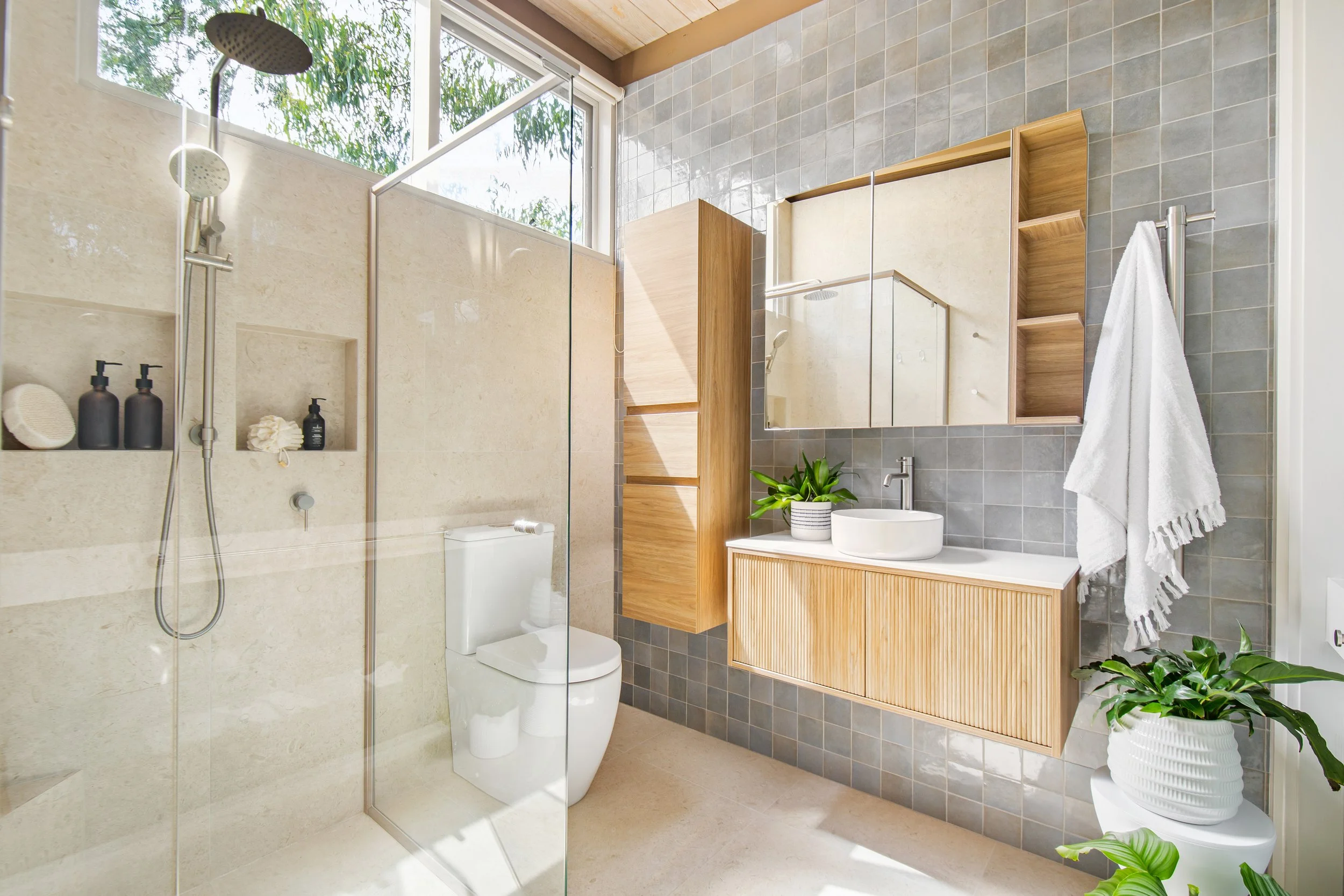 Modern bathroom with beige and gray tiles, a glass shower enclosure, a wall-mounted mirror cabinet, a white vessel sink with a wooden vanity, potted green plants, and a light-filled space with large windows.