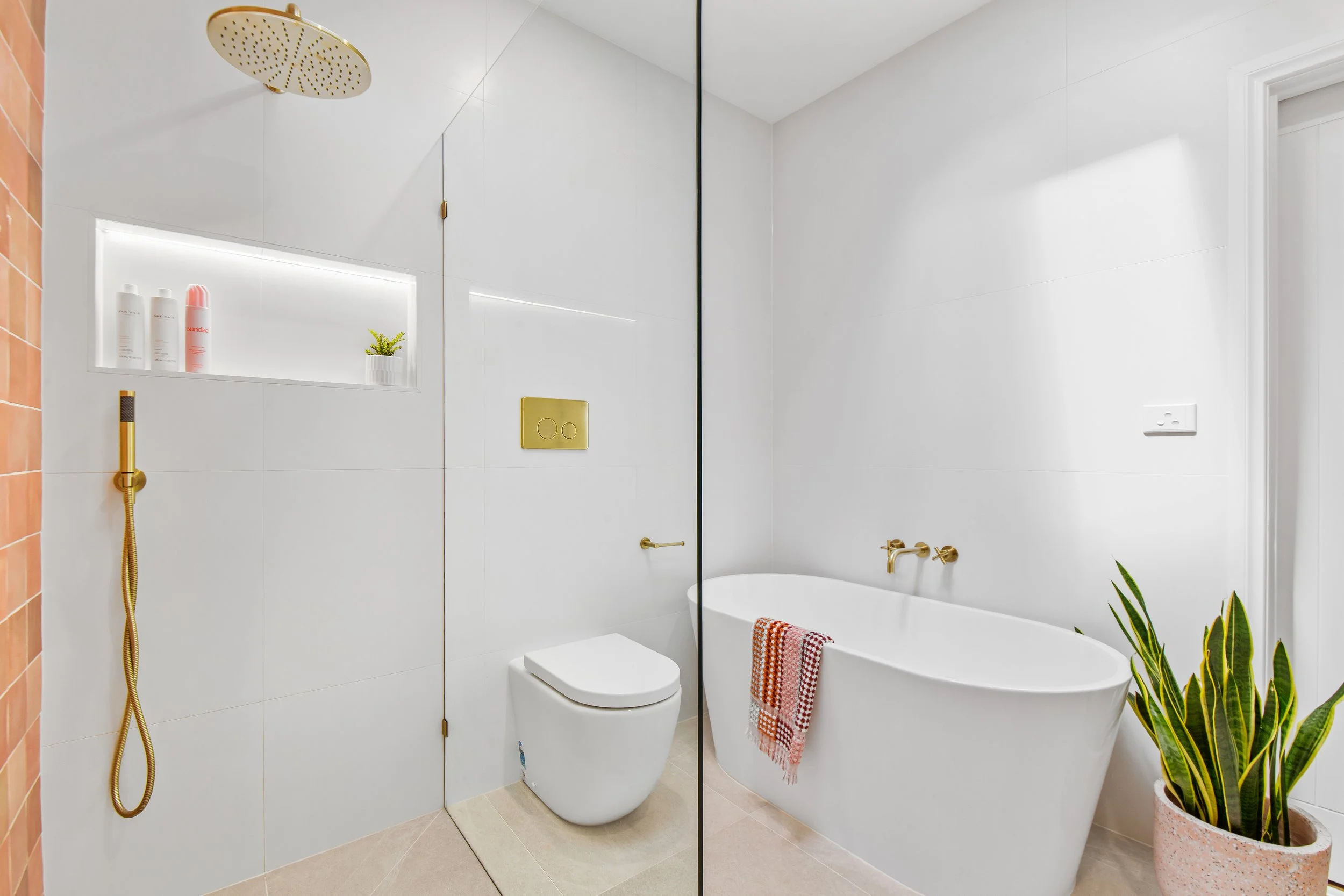 Modern bathroom with white walls, a bathtub with a towel, a wall-mounted gold faucet, a toilet, a showerhead, a niche with bottles and a plant, and a large potted plant.