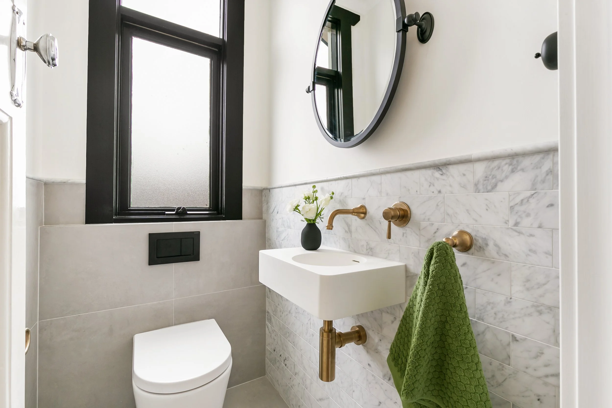 Small modern bathroom with a wall-mounted sink, green towel, round mirror, black-framed window, and brass fixtures.