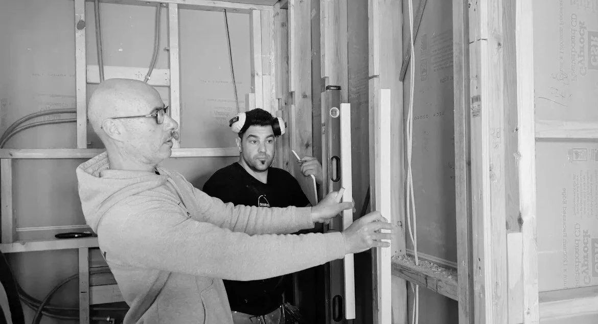 Two men working on construction framing, one demonstrating using a level tool while the other observes, in a partially built room with electrical wiring and insulation.