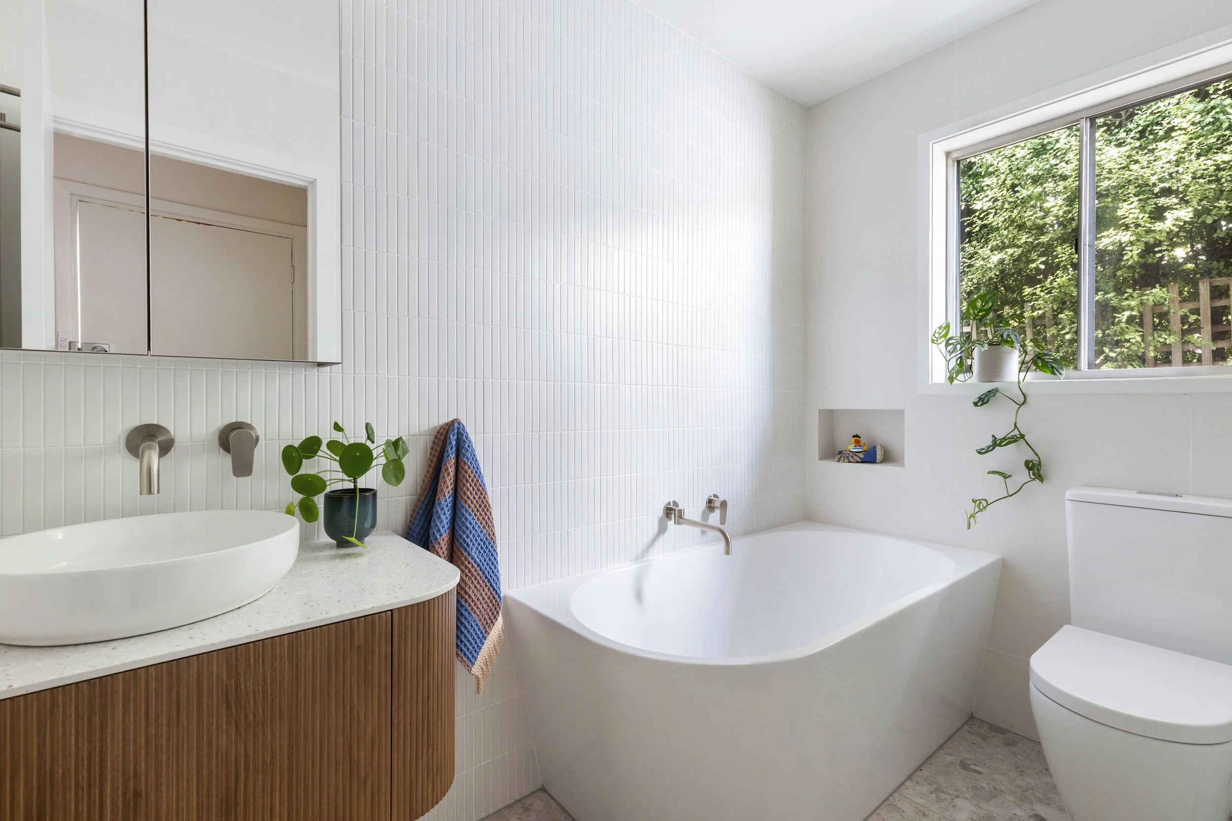 Modern bathroom with white tiled walls, a free-standing bathtub, a wooden vanity with a vessel sink, a green potted plant, and a large window with greenery outside.