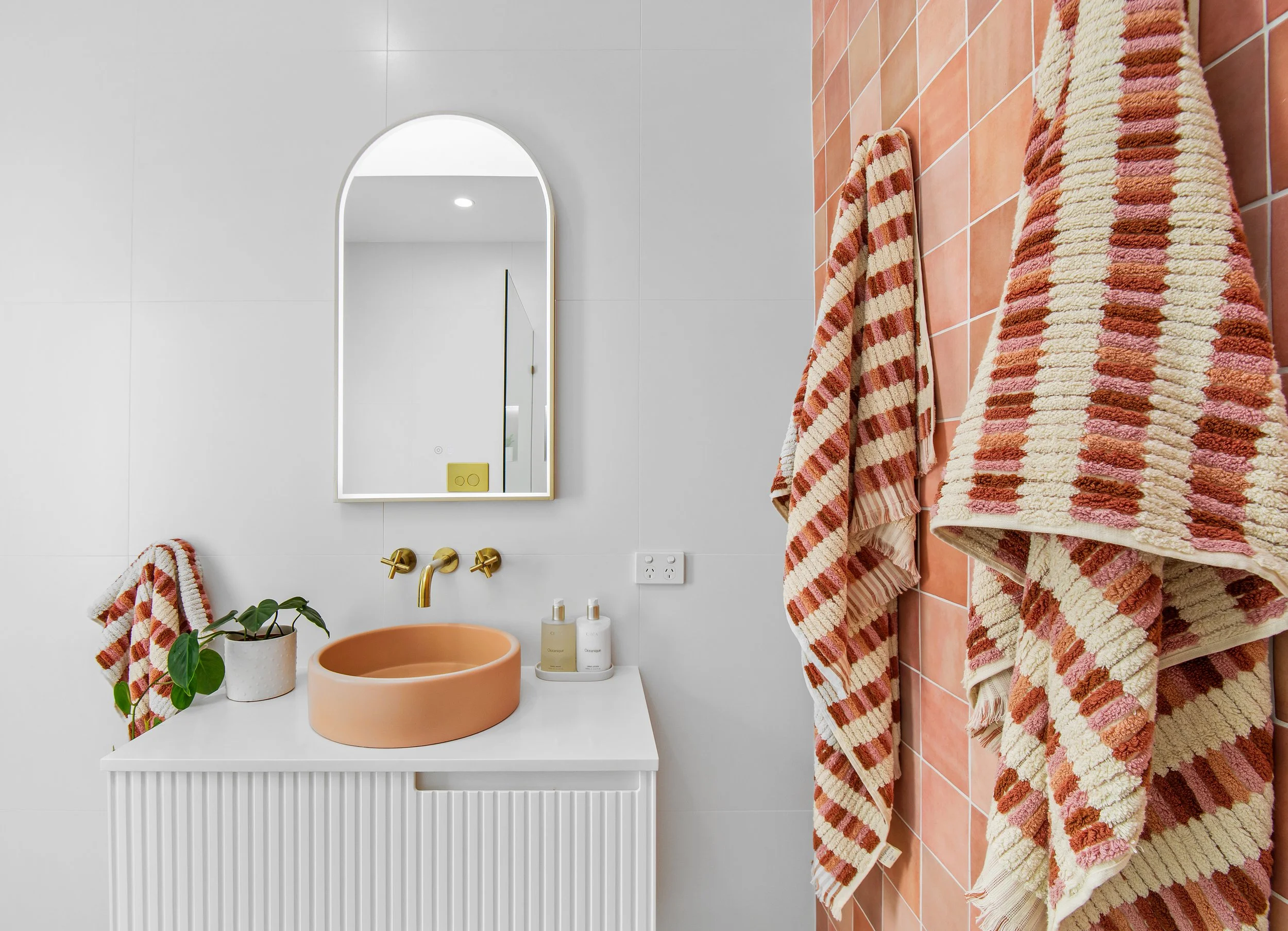 A bathroom with a white wall and a white cabinet with a peach-colored basin sink. Above the sink is a mirror with an arch top. To the right, the wall is tiled with pinkish-orange tiles, and hanging on it are several brightly-colored, striped towels i
