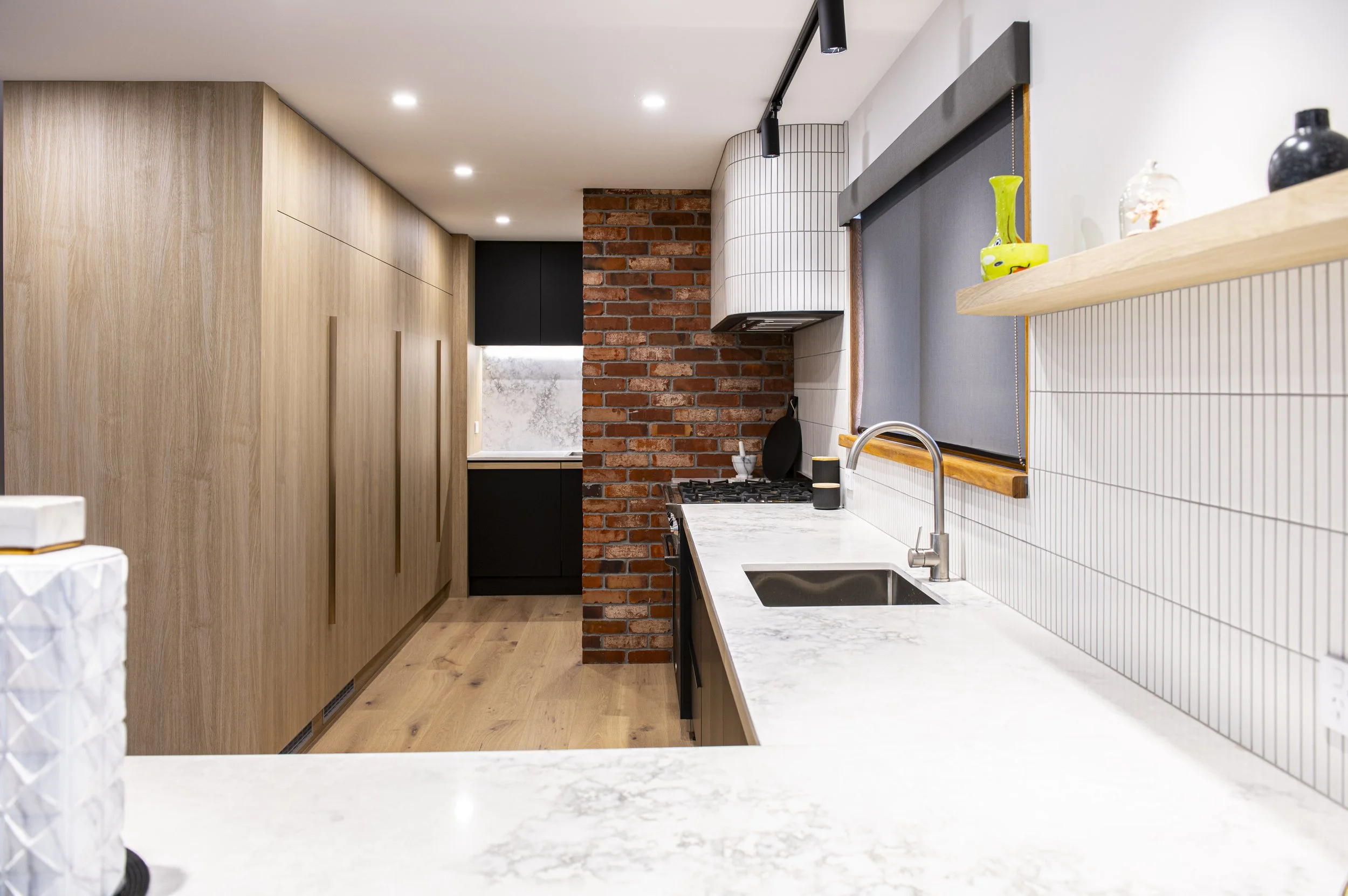 Modern kitchen with wooden cabinets, white marble countertops, brick accent wall, black appliances, and a window with a gray roller blind.