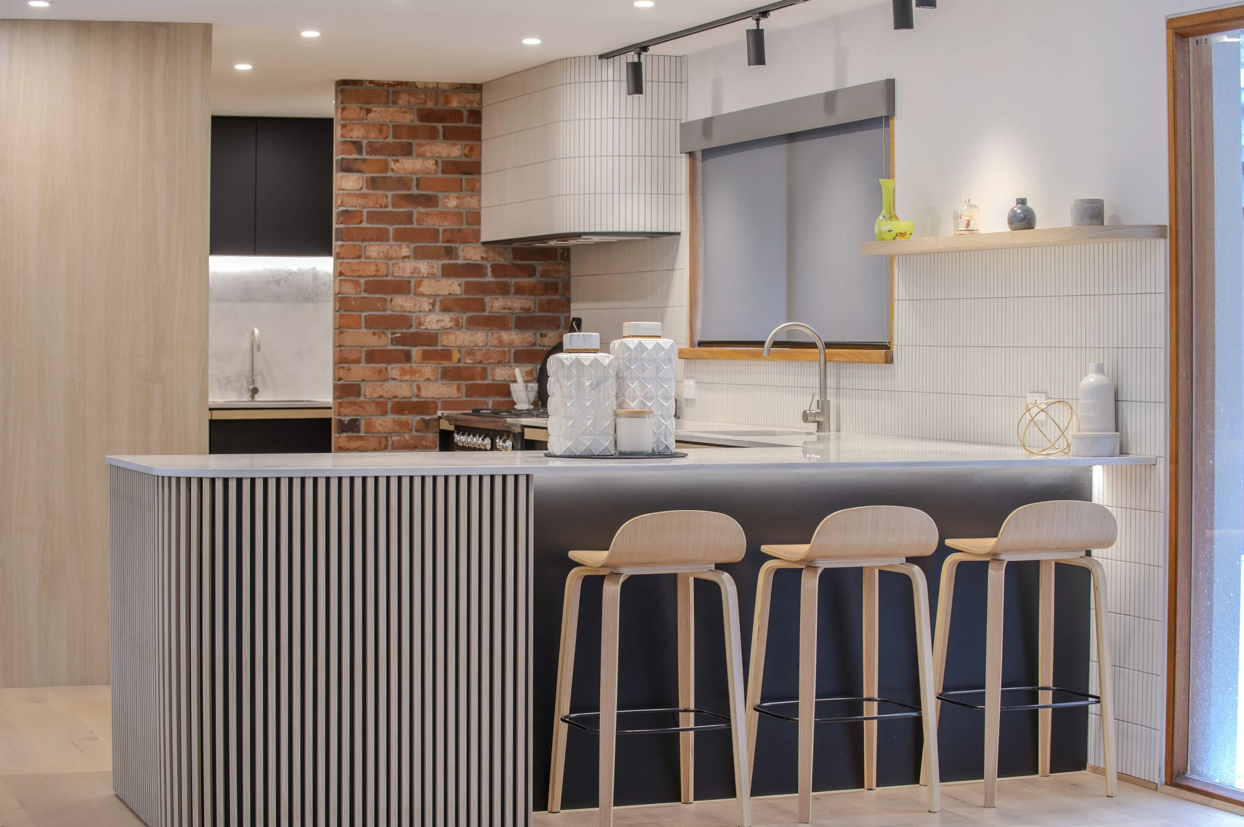 Modern kitchen with white countertop, brick accent wall, beige barstools, wooden accents, and decorative items.