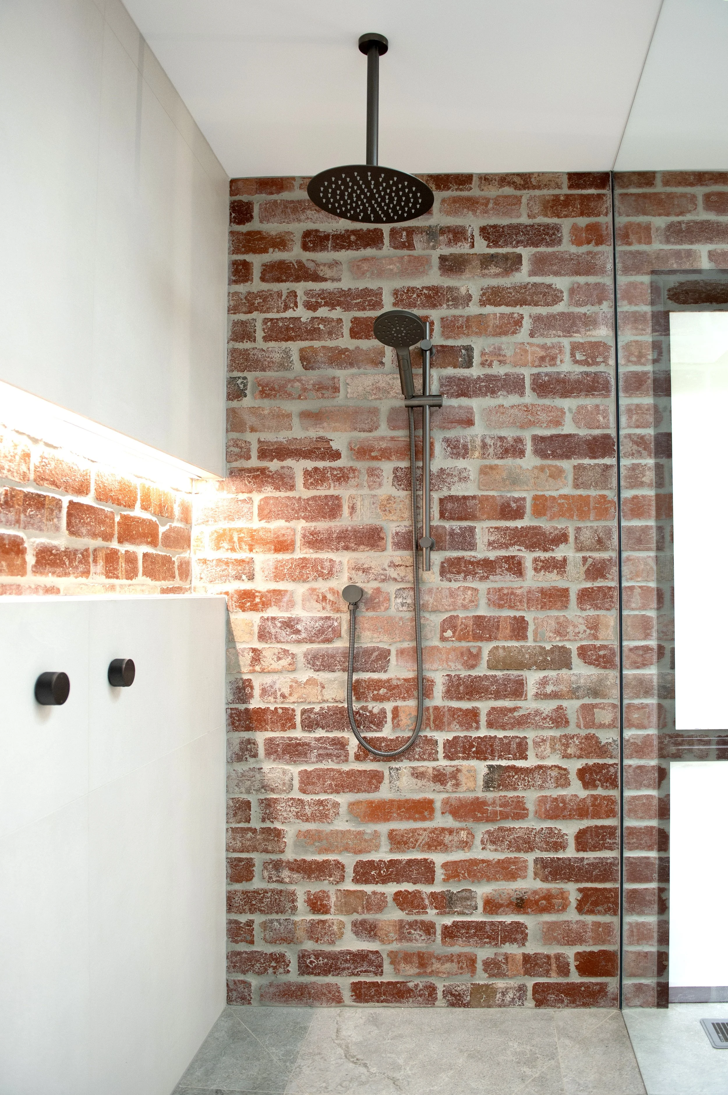 Modern shower with a brick accent wall, black rain showerhead, handheld shower, and white cabinetry.
