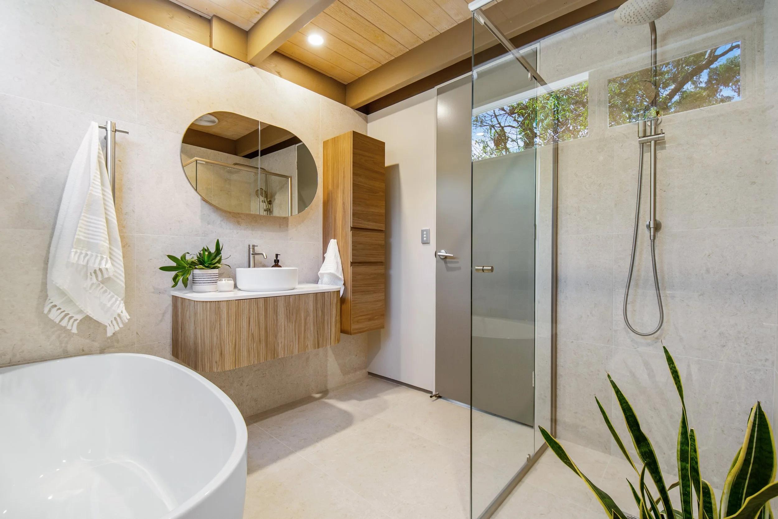 Modern bathroom with a freestanding bathtub, a wooden vanity with a round mirror, and a glass-enclosed shower with a rainfall showerhead. The space features natural wood accents, a potted plant, and natural light coming through a window.