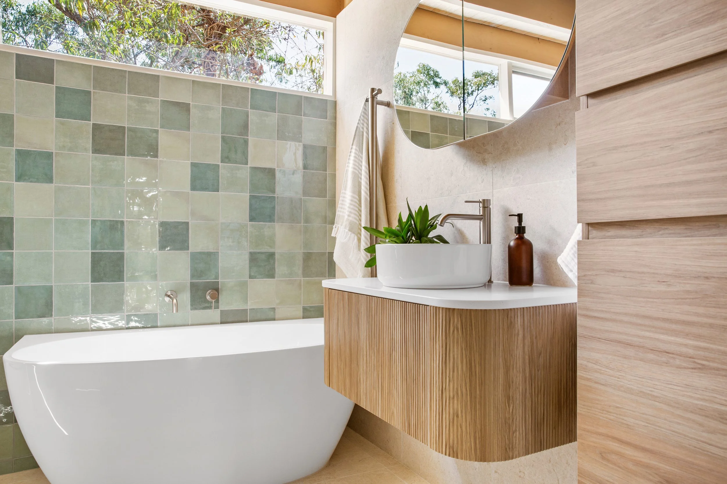 Modern bathroom with a white bathtub, green tiled wall, wooden vanity with a white vessel sink, mirror, and a window with trees outside.