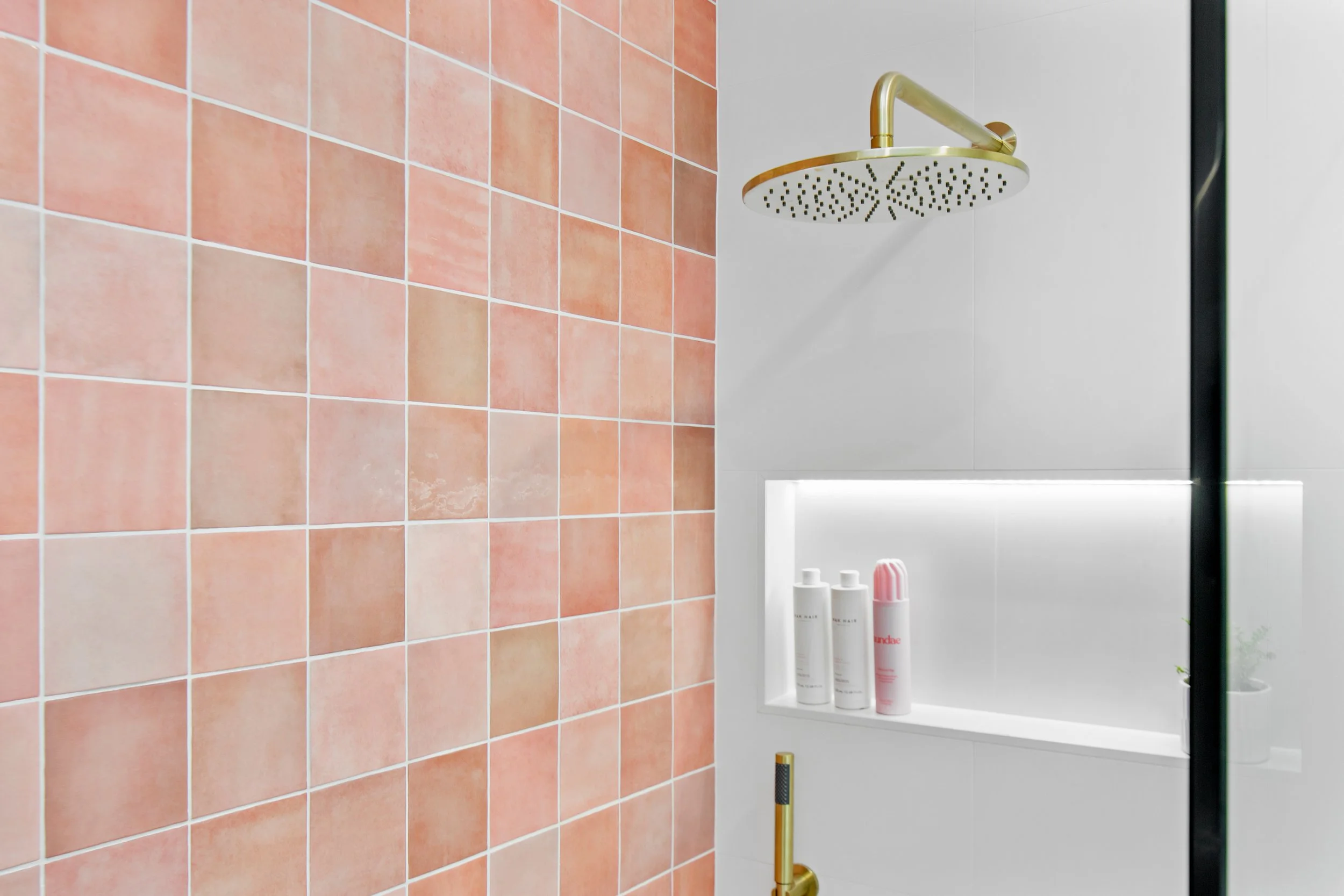 Shower area with pink tiles on one wall, a round gold showerhead, and a white wall with built-in shelf holding bottles of shampoo and a spray can, with a small potted plant nearby.