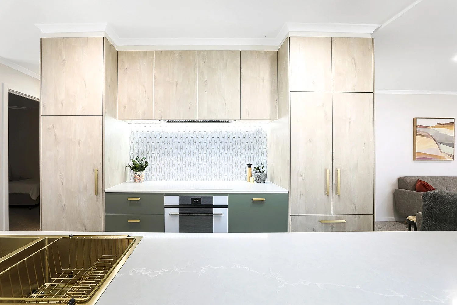 Modern kitchen with light wood cabinets, white countertops, a backsplash with a honeycomb pattern, and a built-in oven. Potted plants and kitchen utensils are placed on the counter. Part of a living room is visible with a gray sofa and wall art.