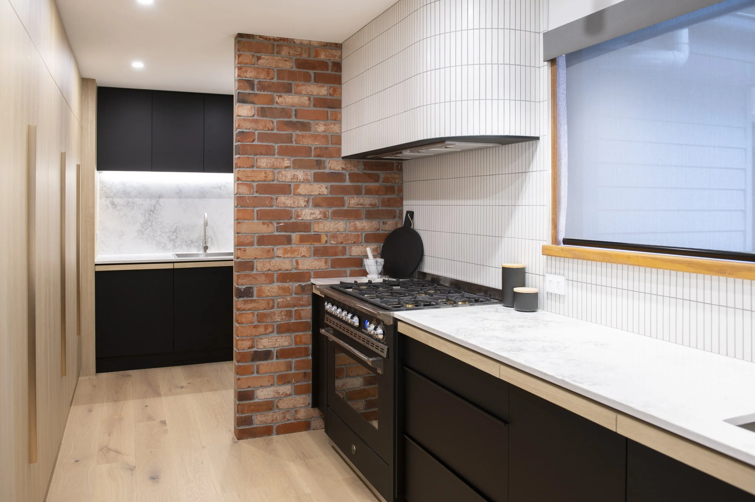Modern kitchen featuring black cabinetry, a brick wall, a white marble countertop, a stove, and a window with a blind.