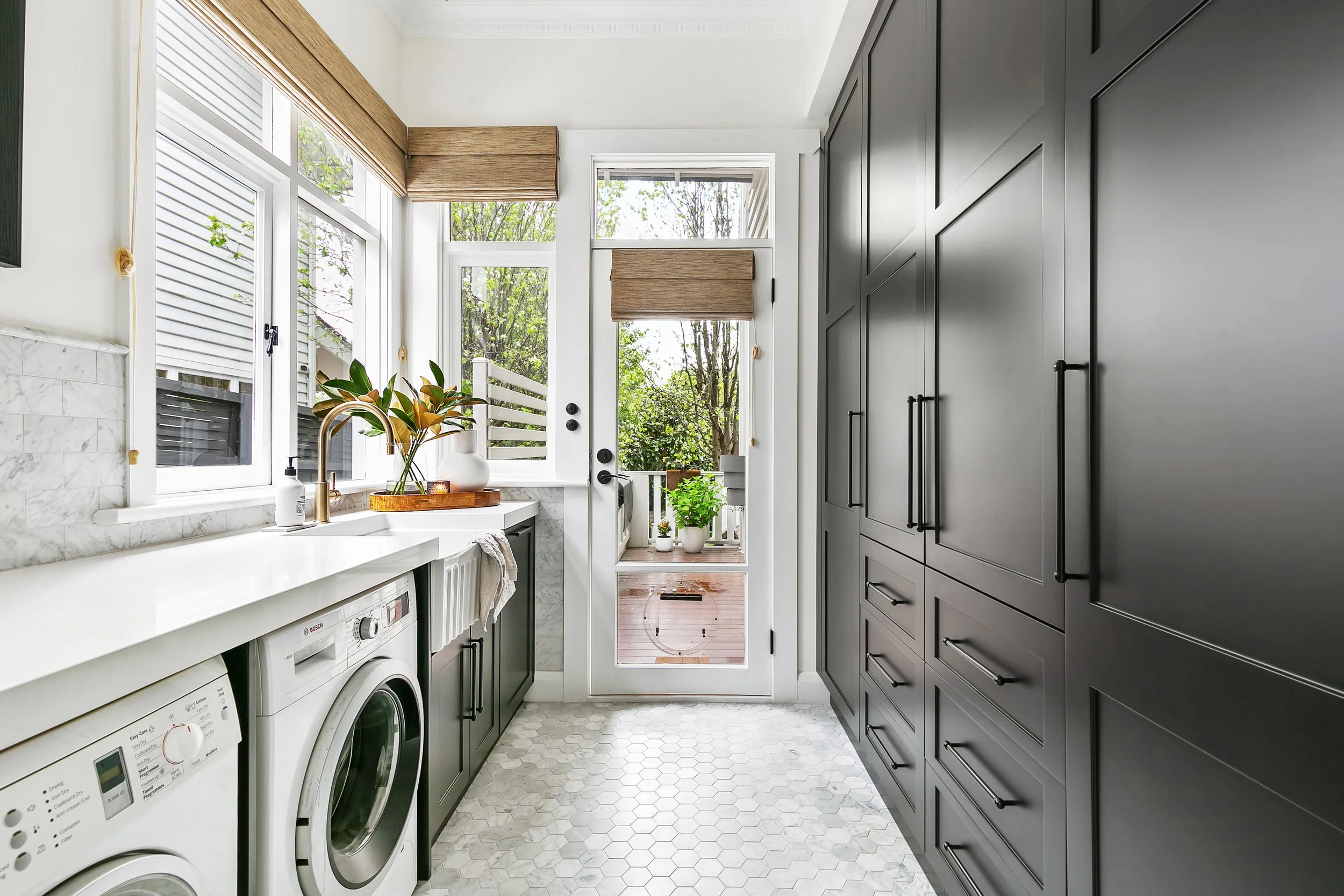 Modern laundry room with white countertops, black cabinets, washer and dryer, and large windows with wooden shades looking onto green trees and a small outdoor space.