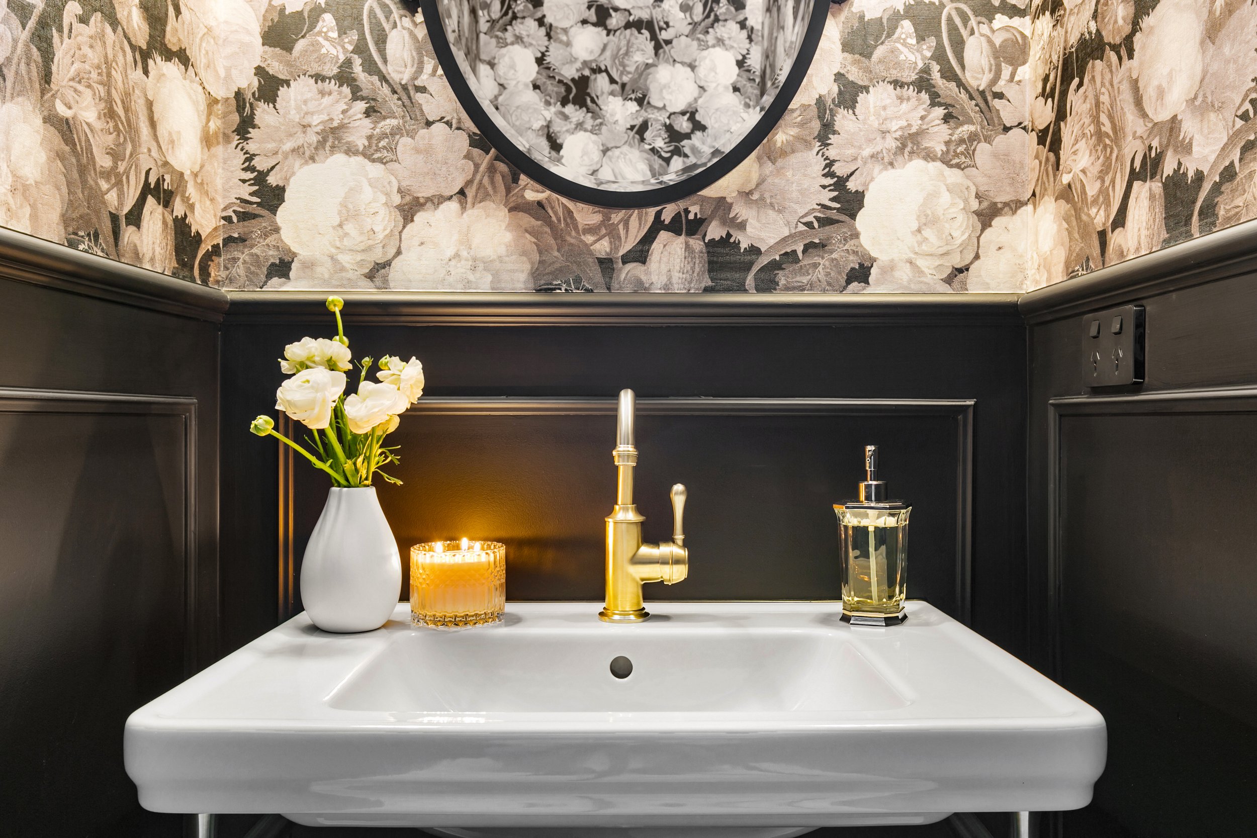 Bathroom sink with a white vase of white flowers on the left, a lit candle in a glass container, a gold faucet, and a soap dispenser on the right, against dark wainscoting and floral wallpaper with a circular mirror above.