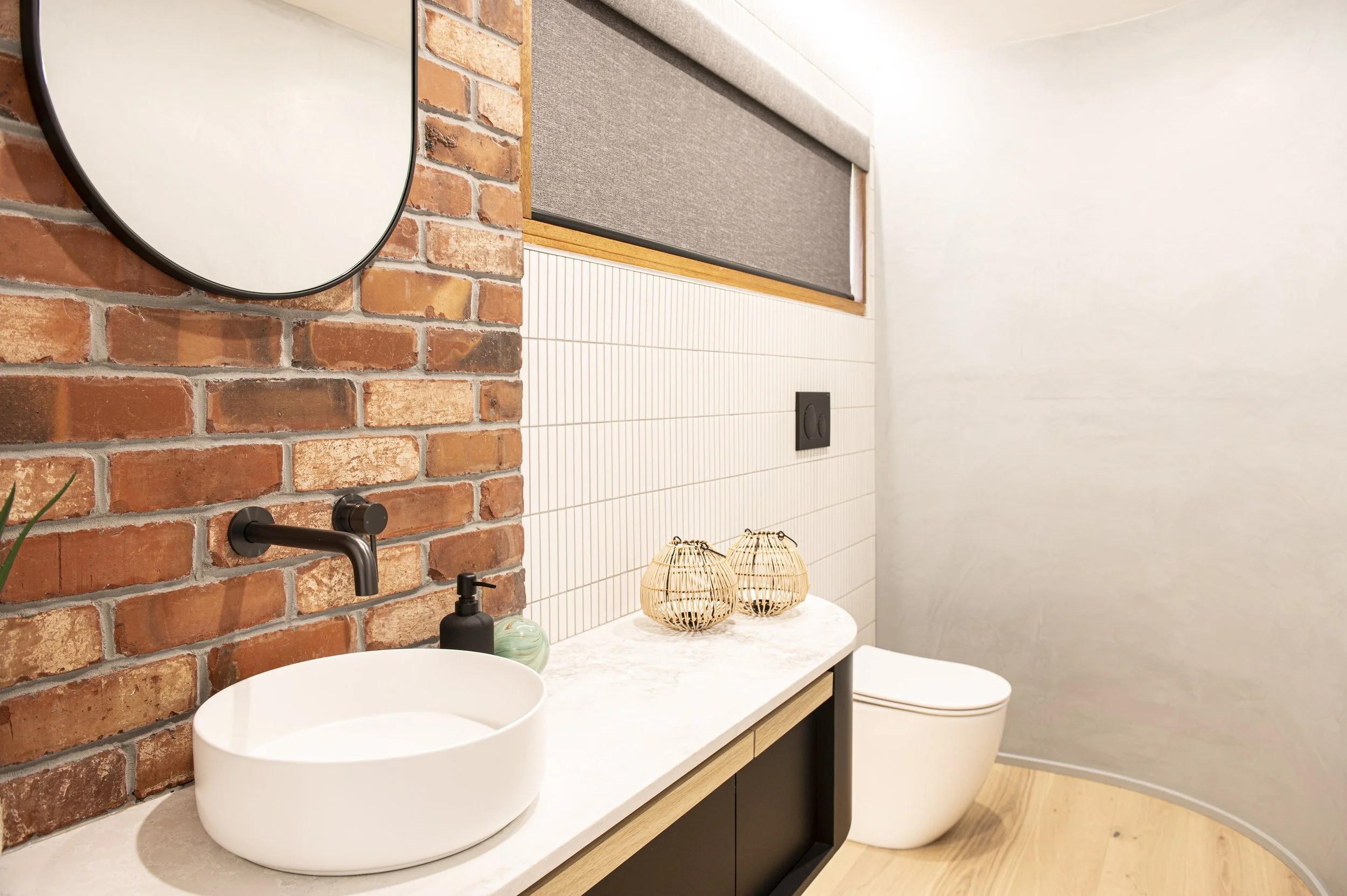 Modern bathroom with a exposed brick wall, a white marble countertop with a vessel sink, a black faucet, a black soap dispenser, and two woven decorative lamps. A white toilet is positioned against a curved wall, with a window covered by a gray rolle