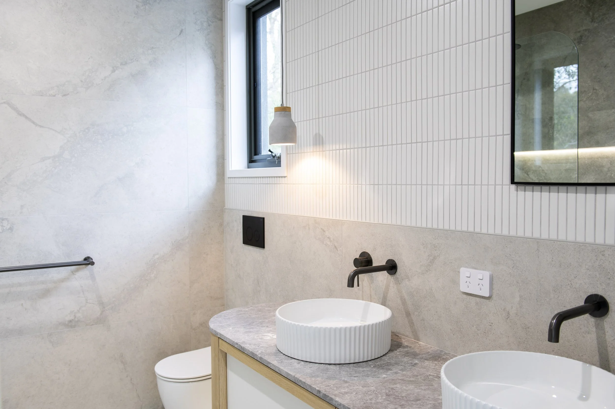 Modern bathroom with double sinks, round vessel basins, black wall-mounted faucets, light-colored stone countertop, textured wall tiles, wall-mounted toilet, small window, and mirror.