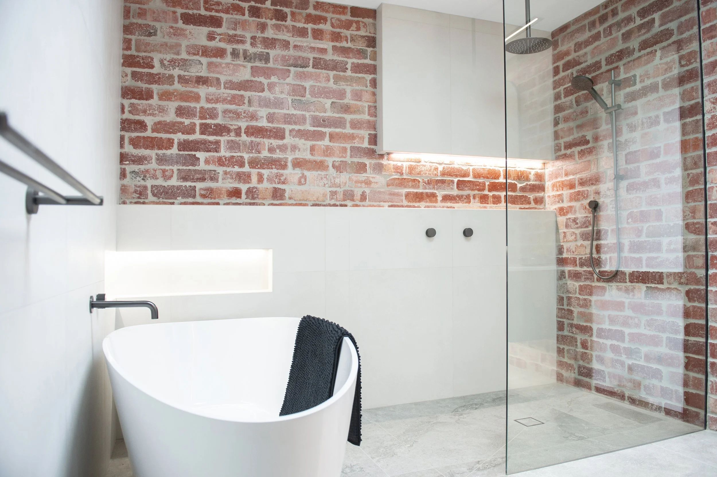 Modern bathroom with a white freestanding bathtub, a brick accent wall, a glass shower enclosure with a rainfall showerhead, and minimalist fixtures.