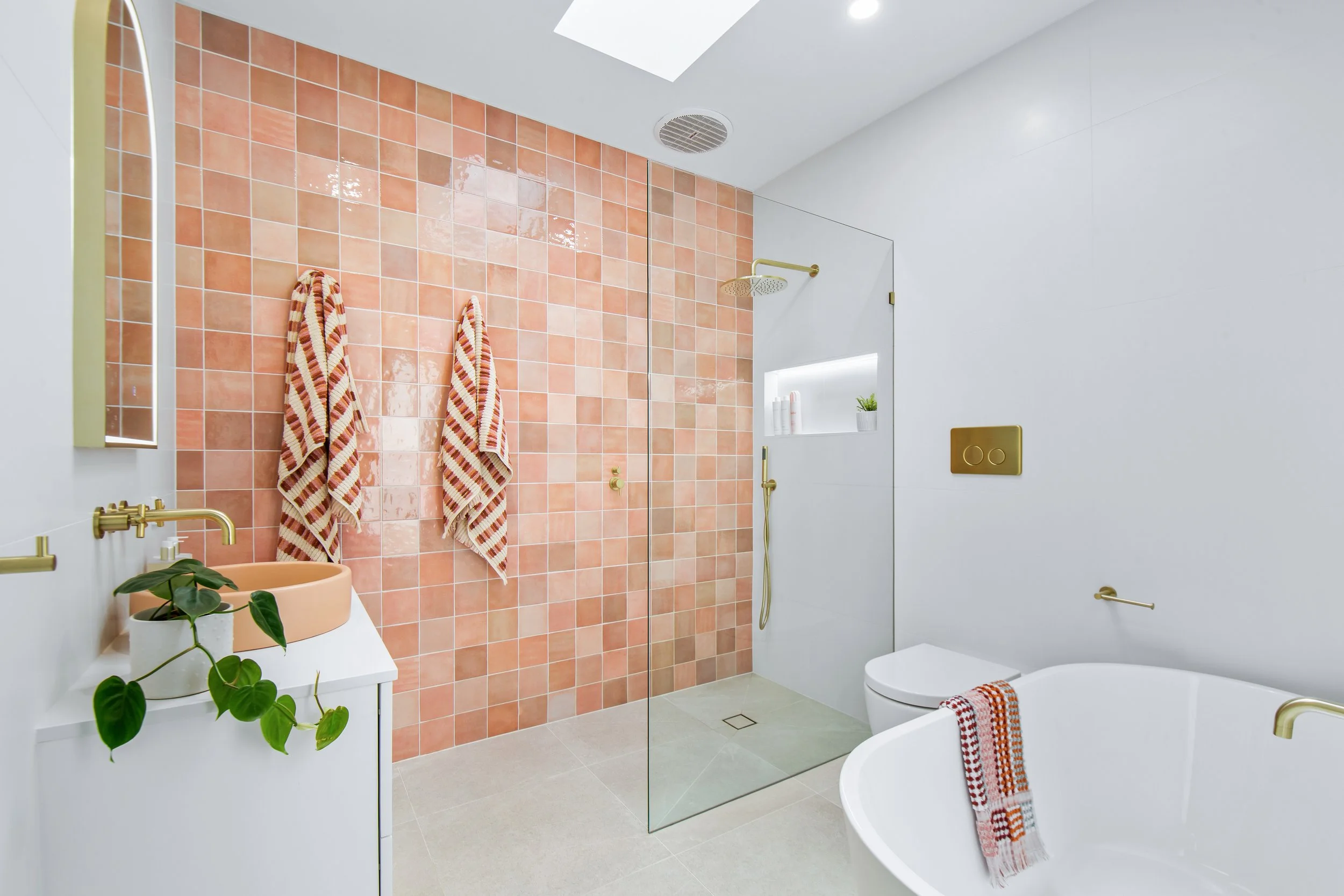 Modern bathroom with peach-colored tile wall in the shower area, a white vanity with a peach sink and green plant, striped towels hanging on hooks, a glass shower partition, a built-in shelf with toiletries and a plant, a white toilet, and a free-sta