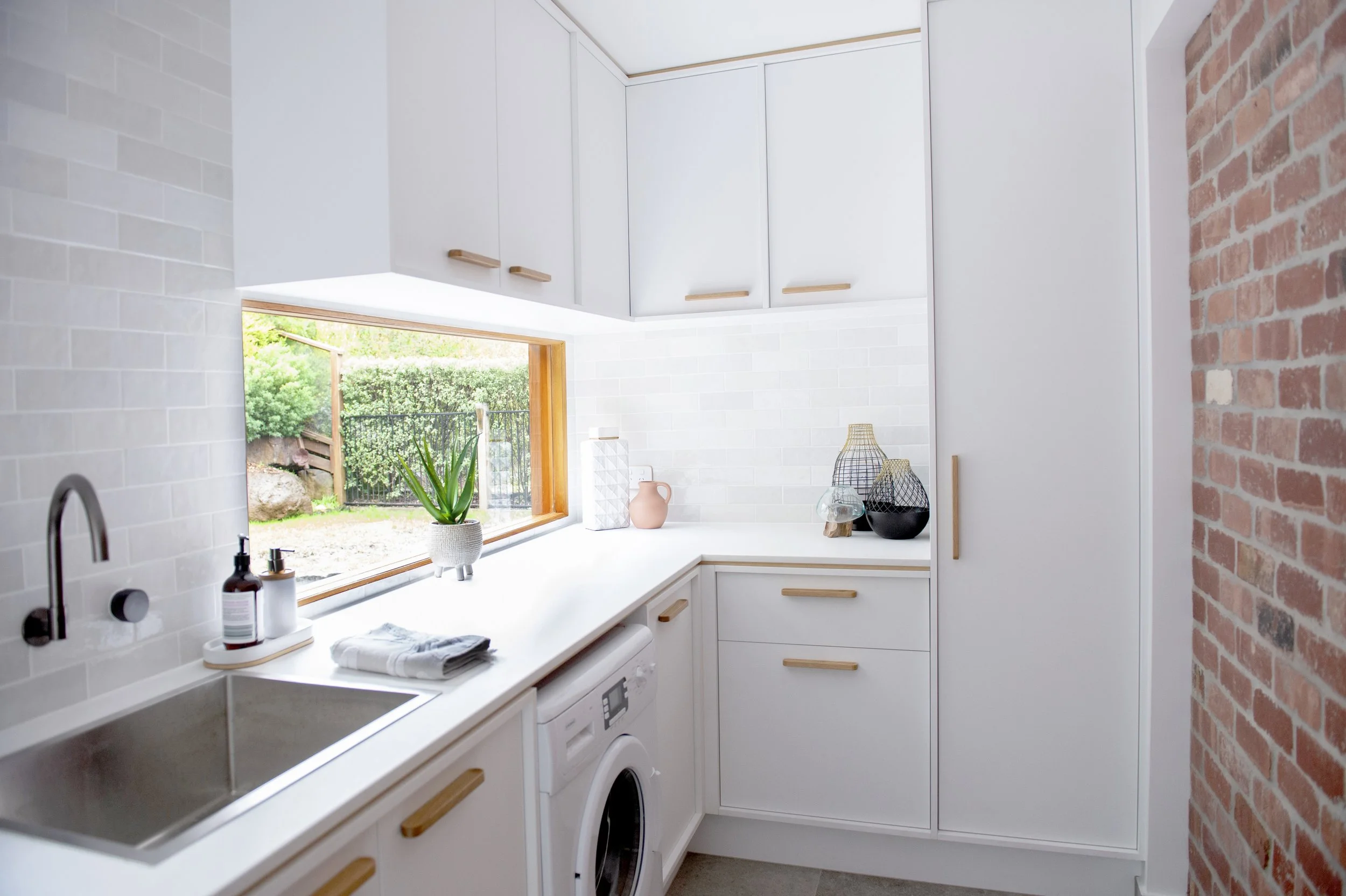 Bright laundry room with white cabinetry, a window with a wooden frame overlooking a yard, a white countertop with a potted plant, decorative vases, and laundry supplies, brick wall on the right, and white tiled backsplash.