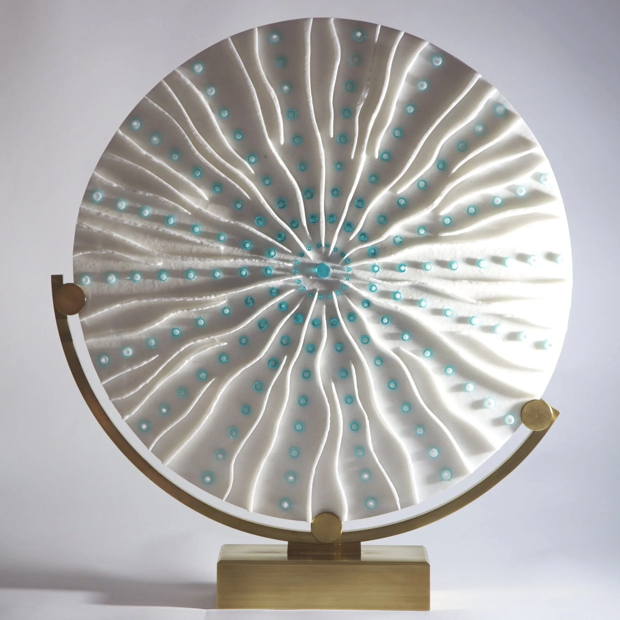 A modern artistic sculpture featuring swirling white fabric-like material with numerous small turquoise circles, mounted on a wooden stand with a circular metallic frame.