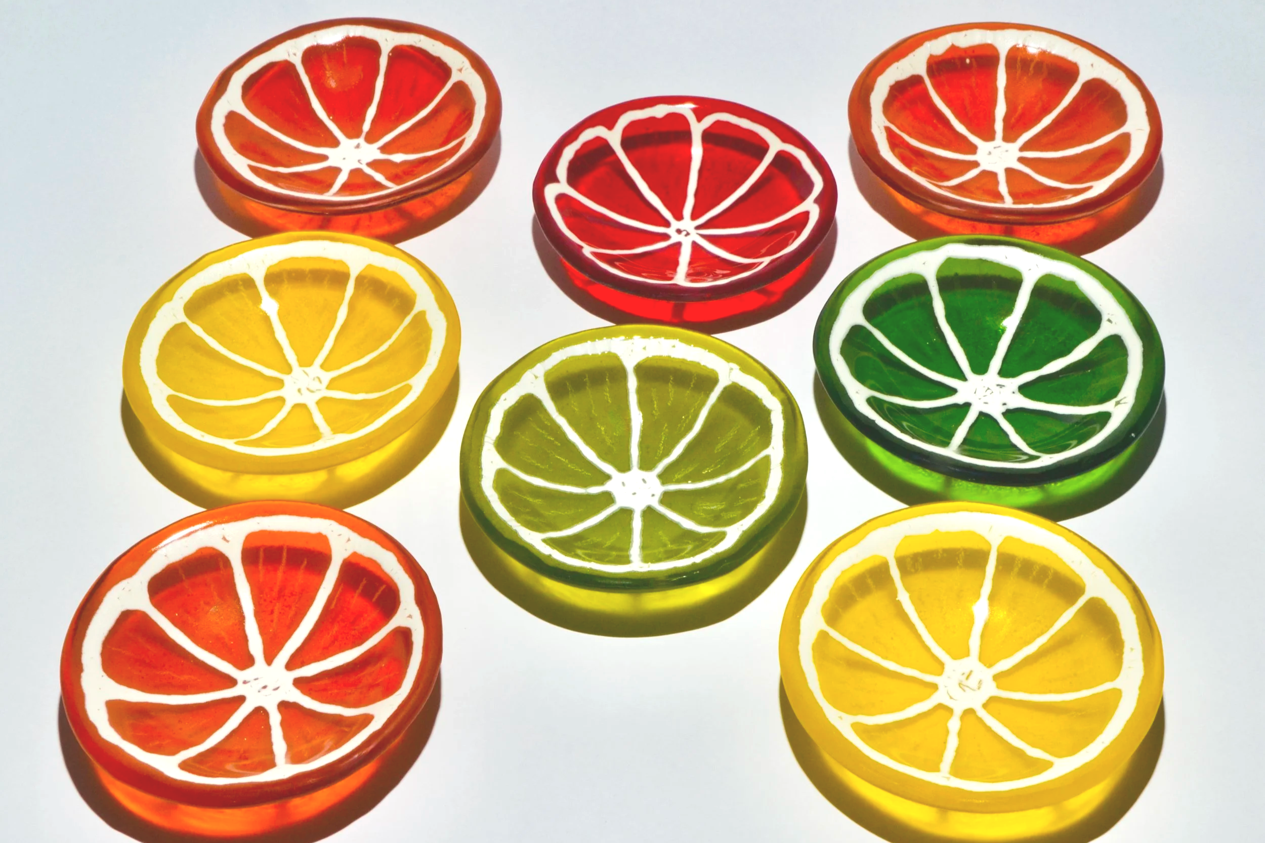 Colorful glass coasters shaped like citrus fruit slices, including orange, lemon, lime, and grapefruit, arranged on a white surface.