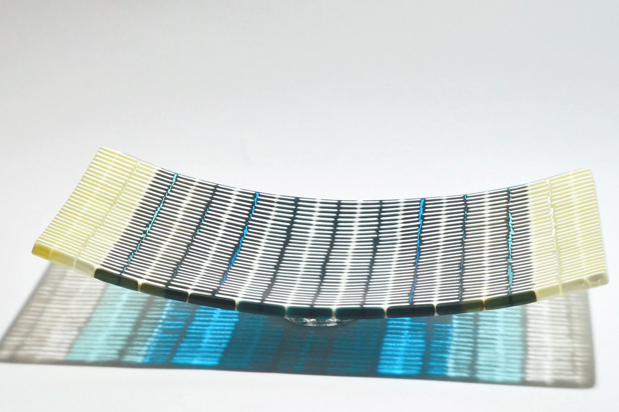 A curved, decorative glass sculpture resembling a multicolored, striped wave, placed on a white surface, casting a vibrant blue and teal shadow below.